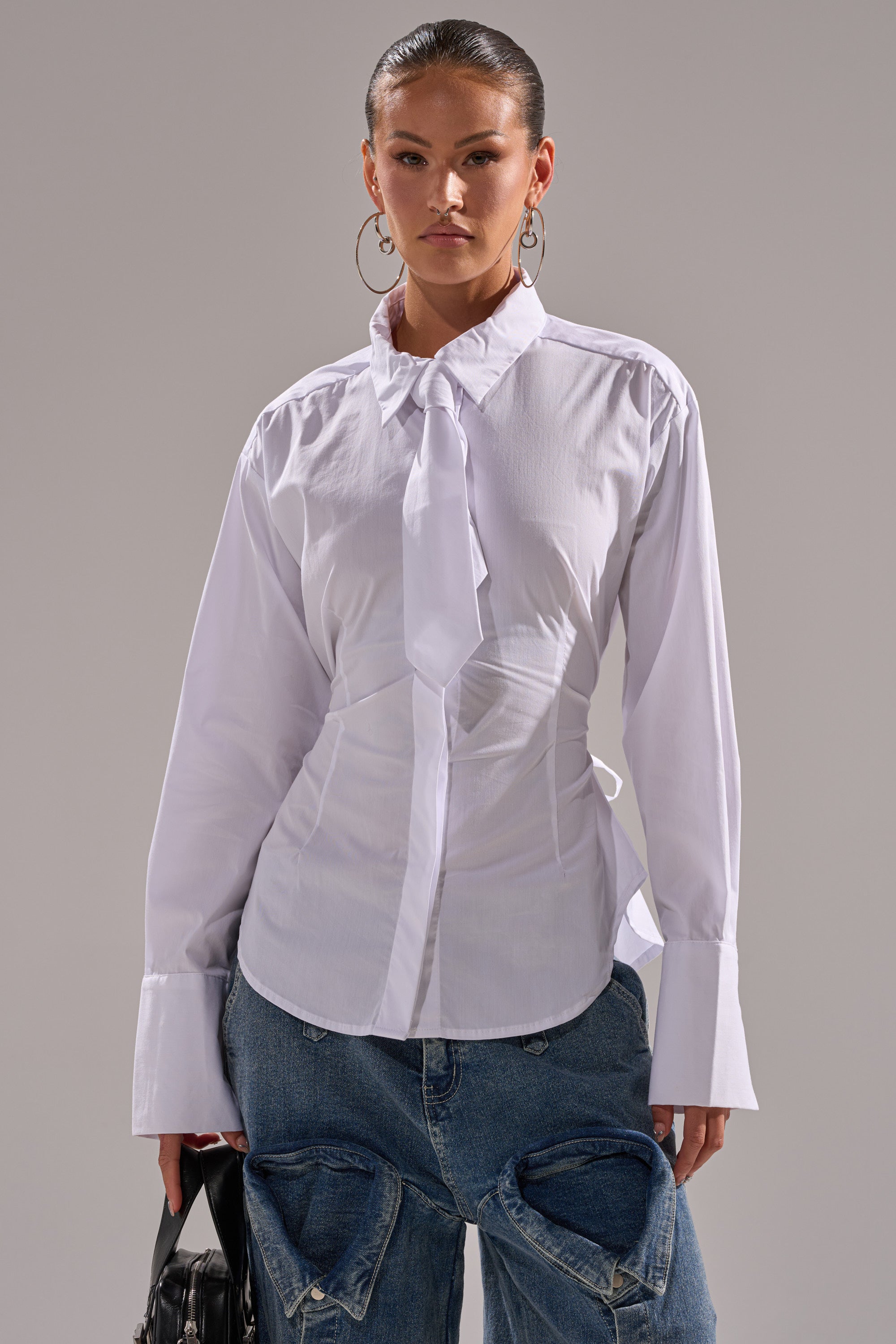 A woman wears the BUSINESS BUTTON DOWN, a white cotton blouse with a necktie and oversized cuffs, styled with baggy blue jeans with large front pockets. She carries a black bag, wears hoop earrings, and has her hair pulled back.