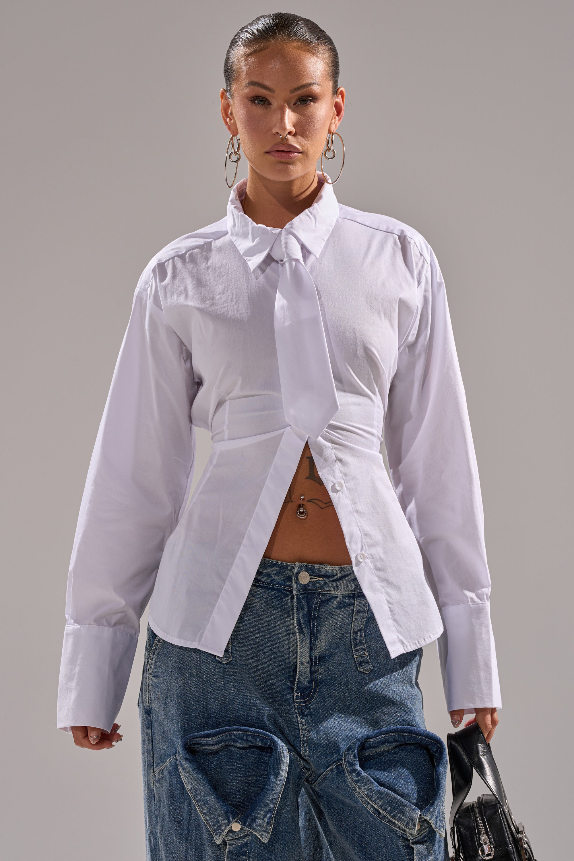 A woman wears the BUSINESS BUTTON DOWN, a white long sleeve cotton shirt with tie detail, paired with blue cutout jeans. She stands against a plain background, holding a small black handbag, with slicked-back hair and hoop earrings.