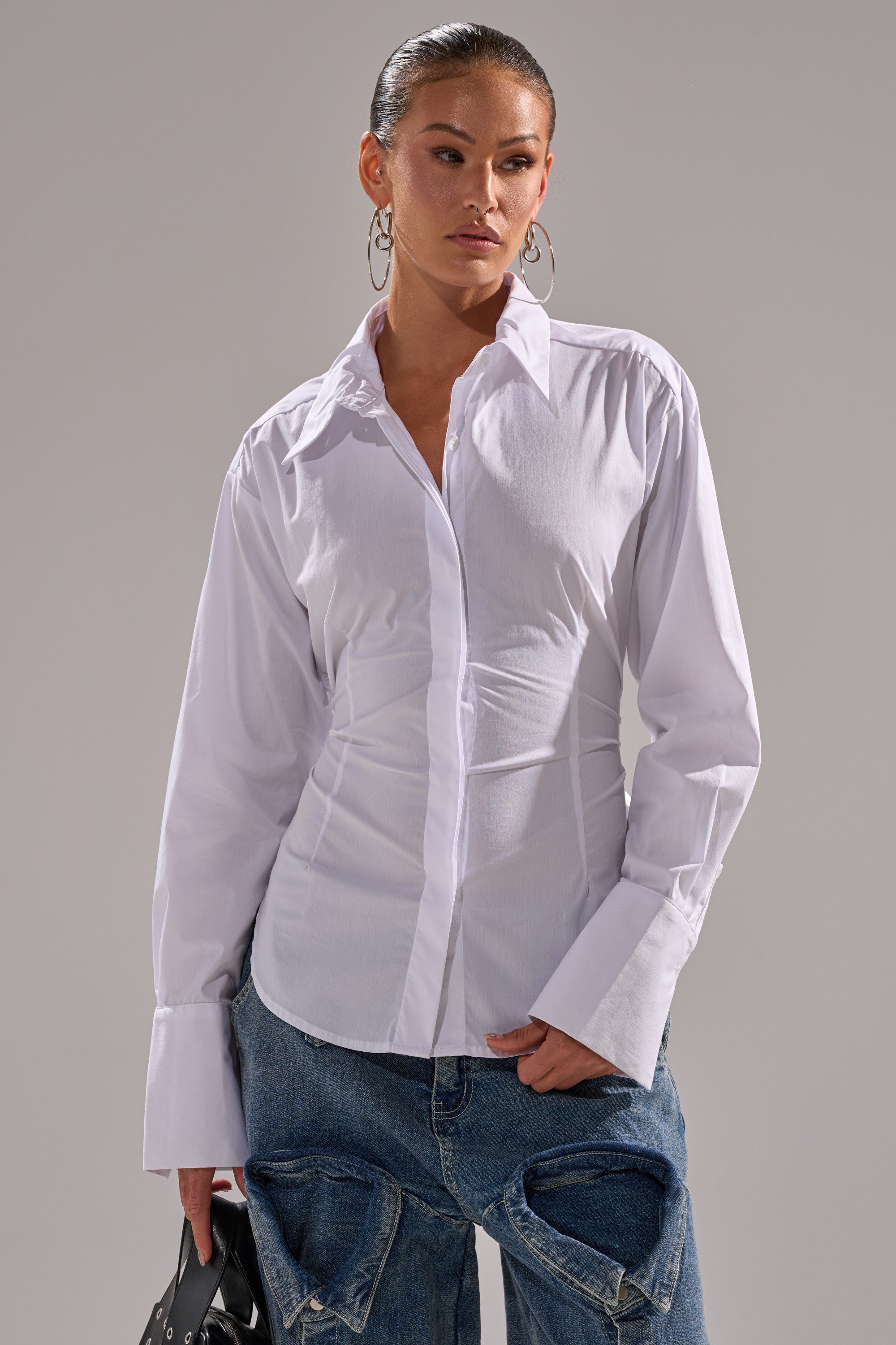 A woman wears the BUSINESS BUTTON DOWN, a crisp white cotton poplin top with oversized cuffs, paired with loose blue jeans and a black bag. She accessorizes with large hoop earrings and has her hair slicked back.