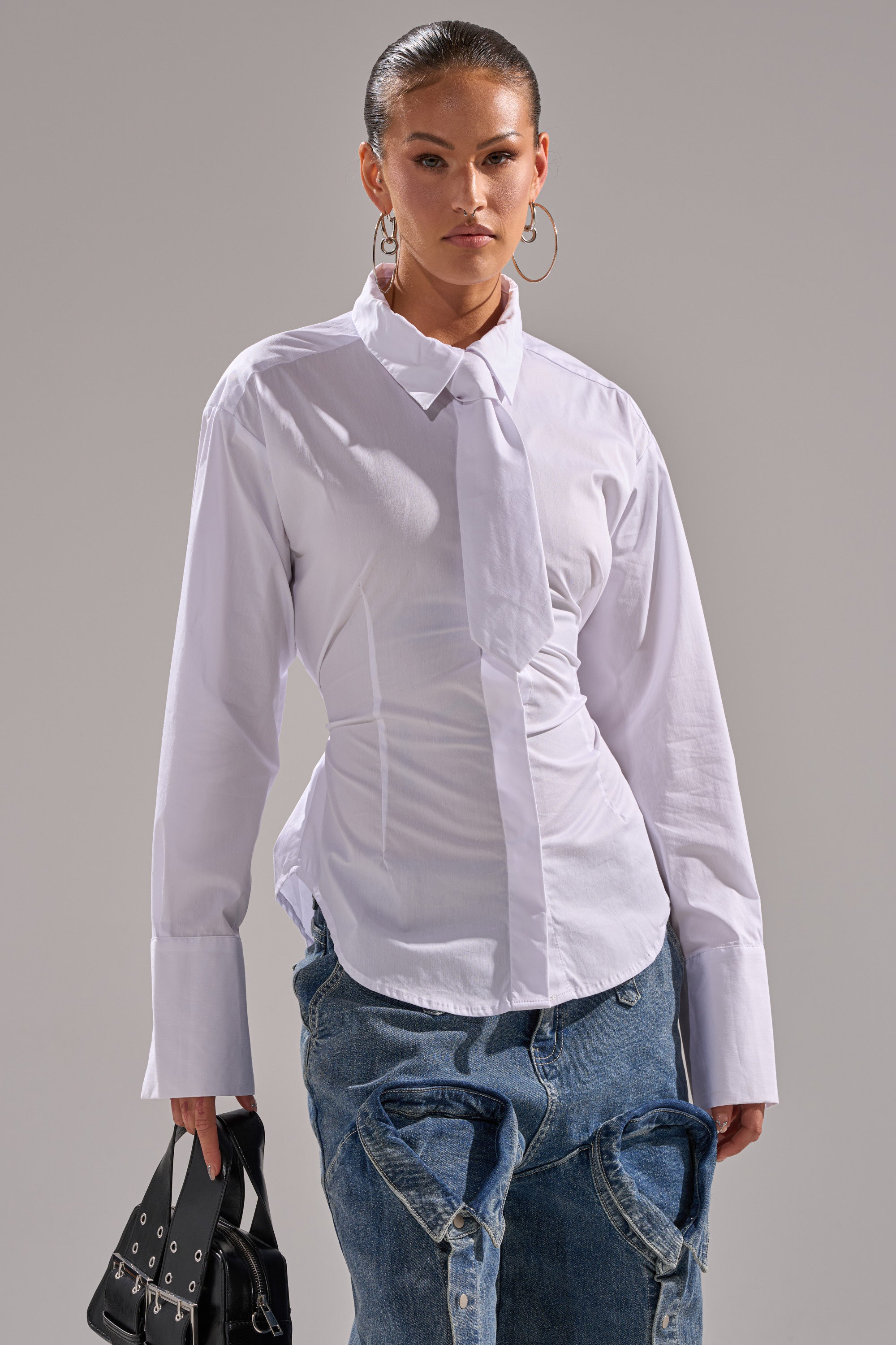 A woman stands confidently in the BUSINESS BUTTON DOWN, a crisp white poplin top with an attached necktie, paired with blue jeans featuring a bold, exaggerated waistline. She completes the look with a small black handbag.