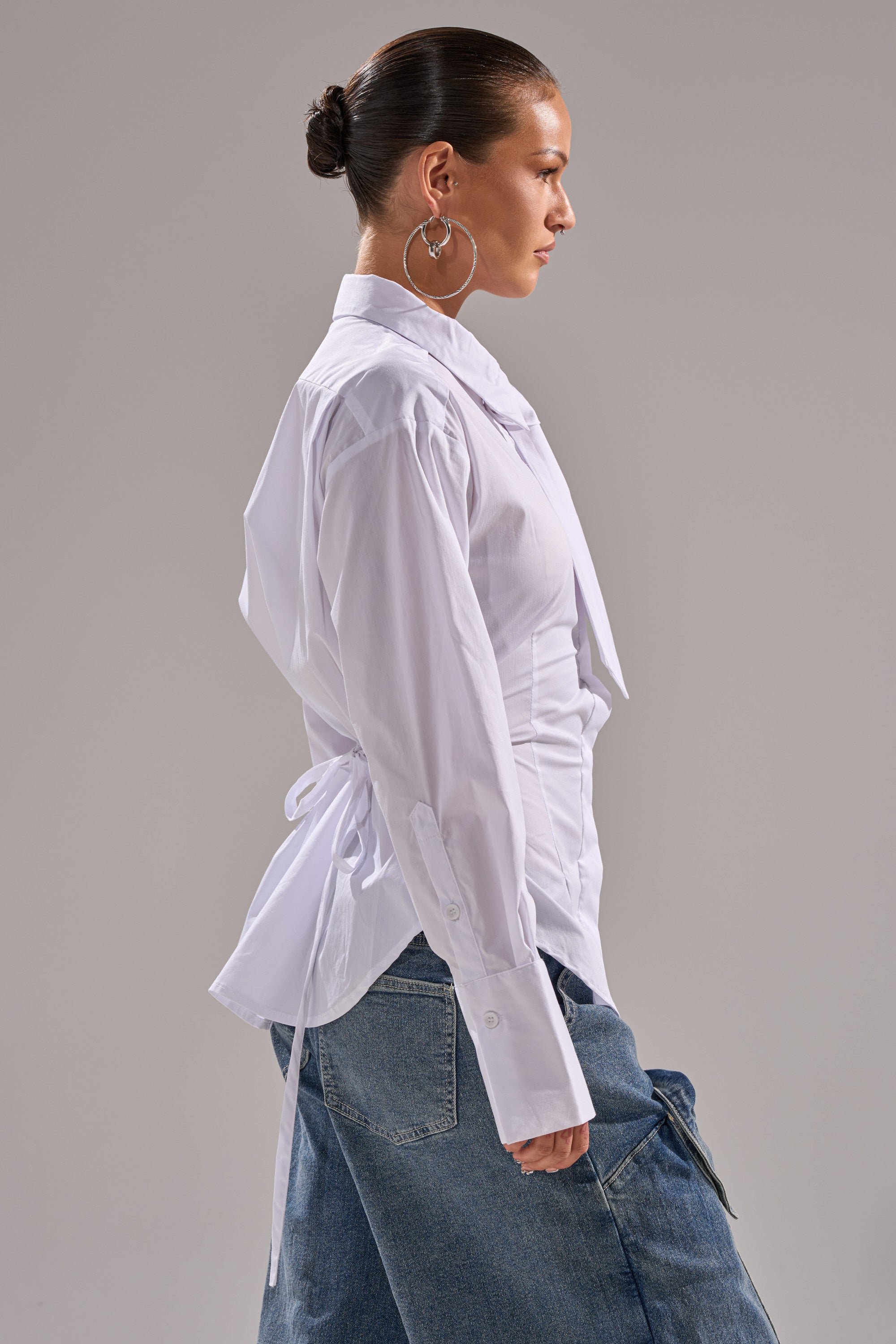 A woman with slicked-back hair in a bun models the BUSINESS BUTTON DOWN, a fitted white blouse with back tie details, paired with loose blue jeans. She stands in profile against a plain gray backdrop.