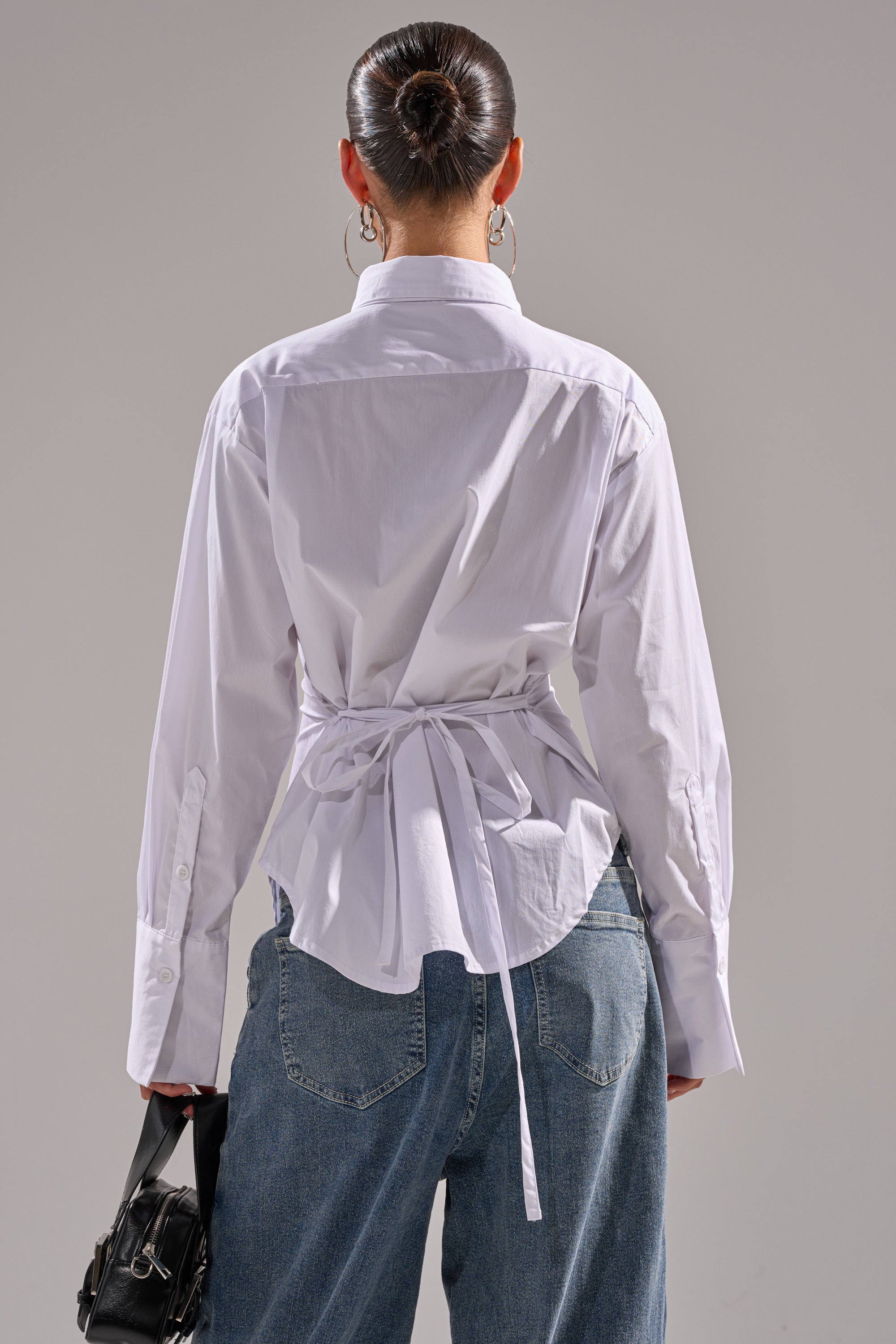 A person stands with their back to the camera, wearing the BUSINESS BUTTON DOWN white cotton blouse tied at the back and loose blue jeans. Hair is in a neat bun. They hold a black handbag in their left hand, set against a plain, neutral background.