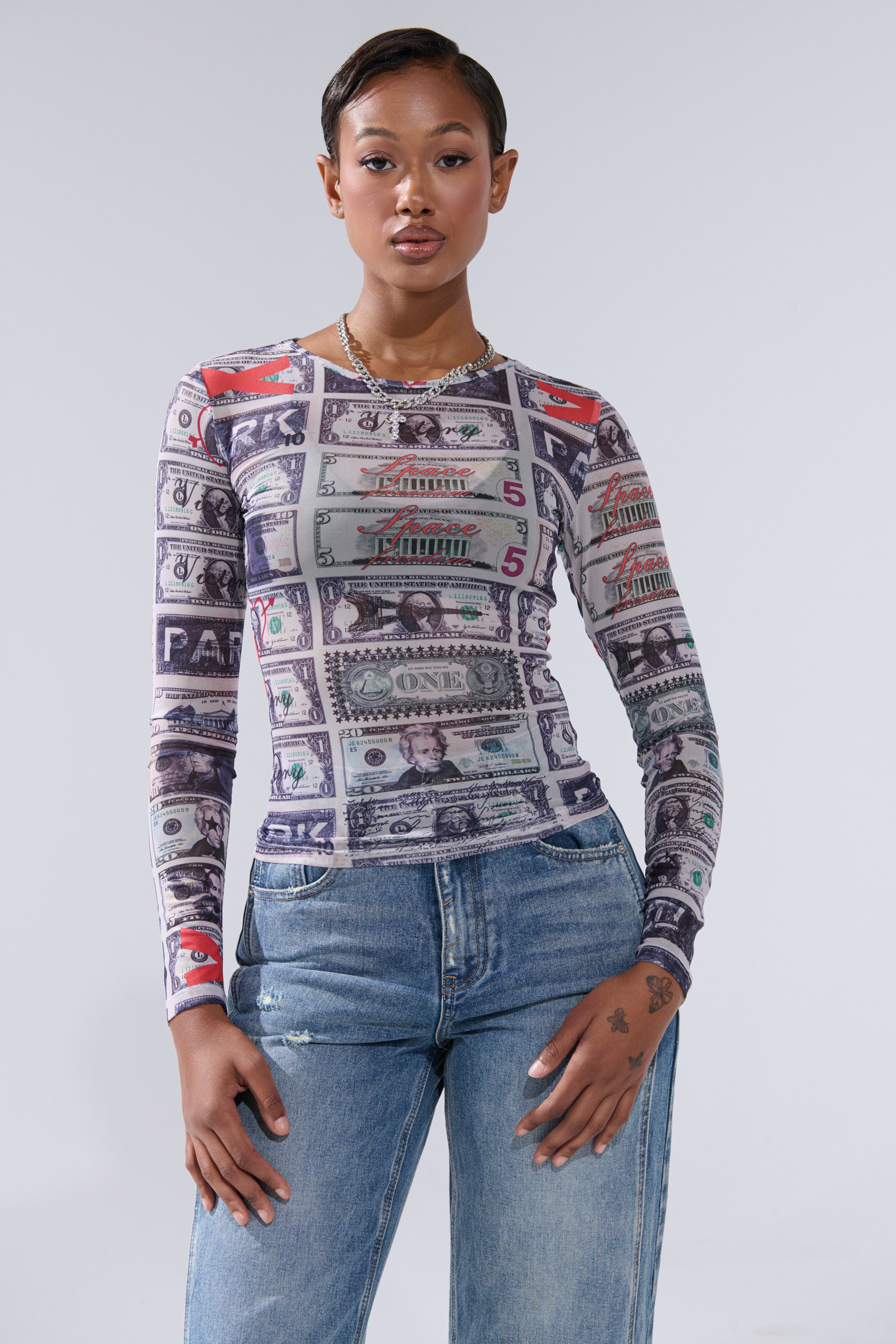 A woman with short hair confidently models the GIMME THE MONEY MESH LONG SLEEVE, featuring dollar sign graphics, paired with blue jeans against a plain light gray background.