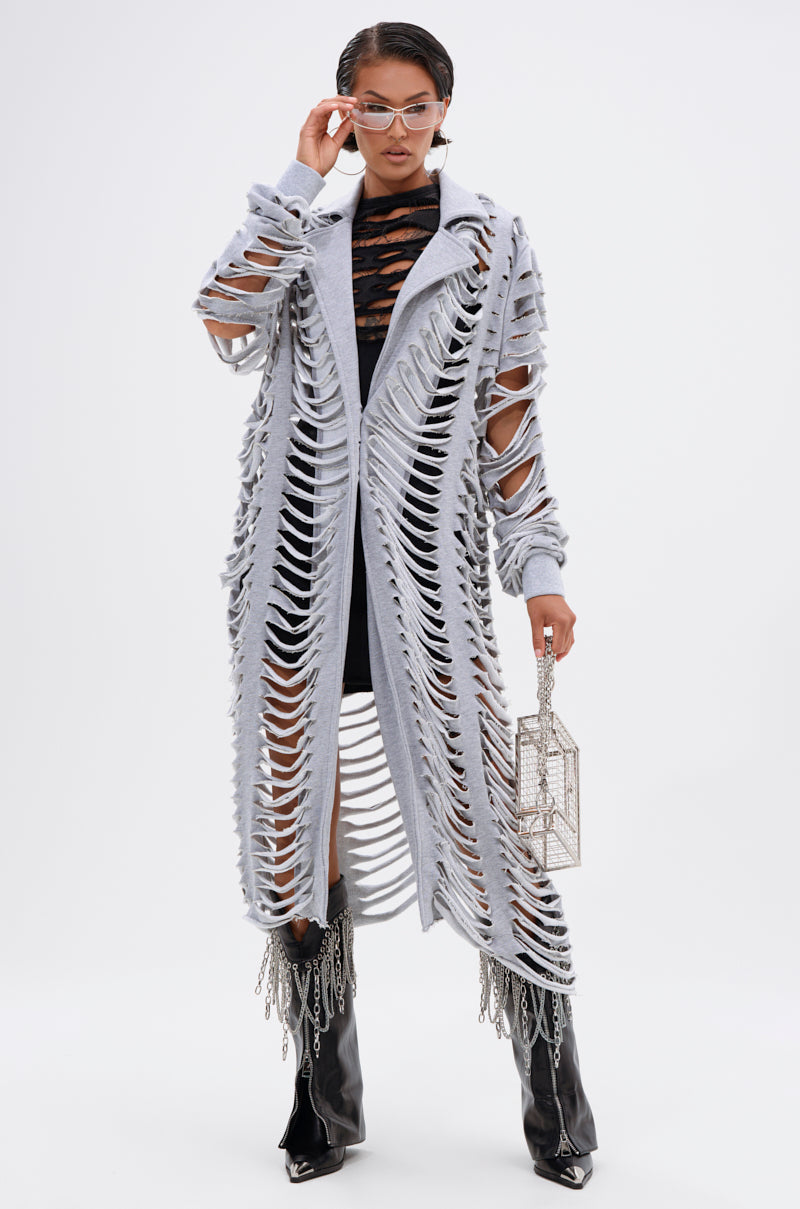 A person stands against a plain white background wearing the CUT IT OUT FRENCH TERRY TRENCH over a black outfit, styled with a geometric silver handbag, silver sunglasses, and black heeled fringe boots. 0