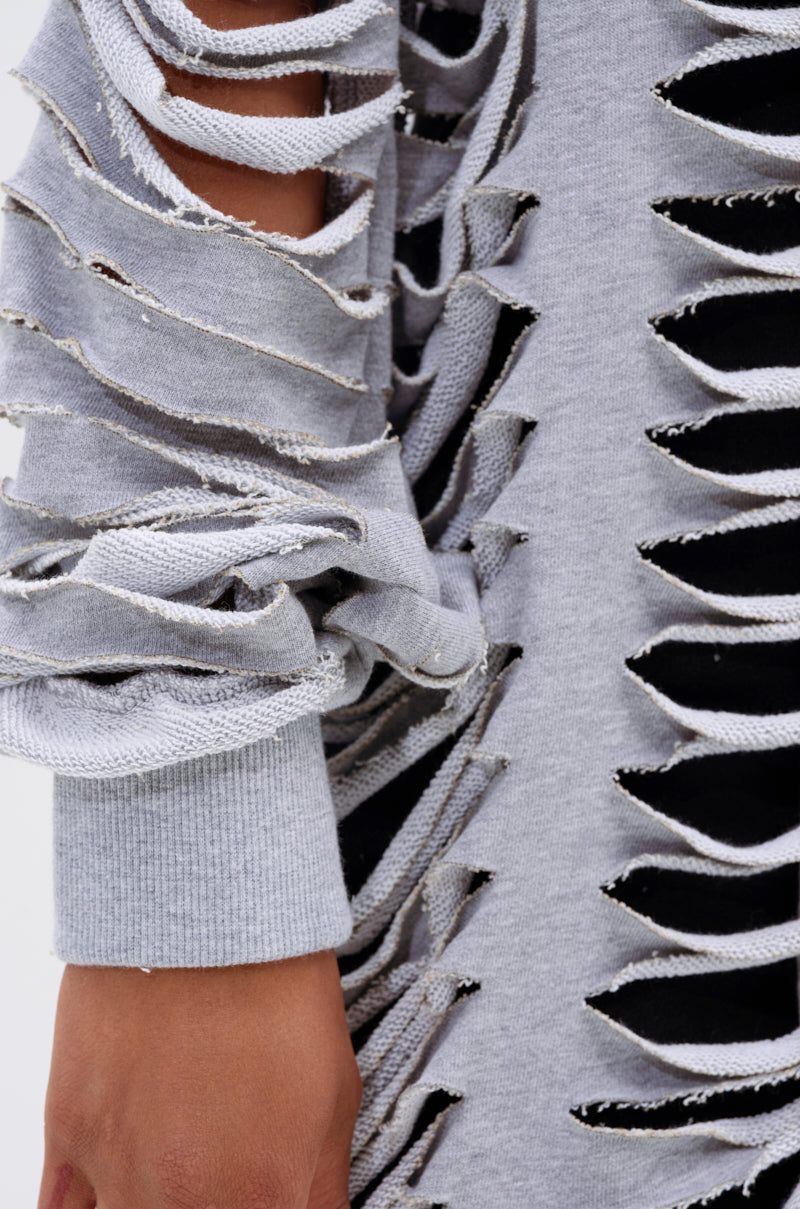 A close-up of a person's arm wearing the CUT IT OUT FRENCH TERRY TRENCH—a light gray sweater with multiple distressed horizontal slits revealing black fabric beneath. The sleeve is scrunched at the wrist, showing part of the hand. 9