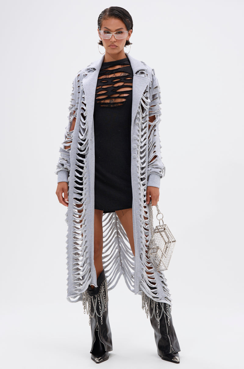 A woman in sunglasses, a black dress, the CUT IT OUT FRENCH TERRY TRENCH in light gray, black pointed boots with silver accents, and holding a geometric beaded handbag stands against a plain white background. 1