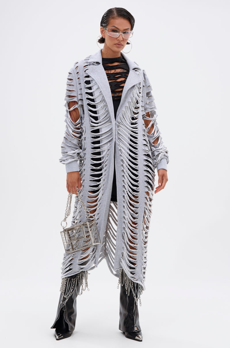 A person stands confidently in a CUT IT OUT FRENCH TERRY TRENCH over a black outfit, paired with black patent boots featuring chains, clear glasses, and holding a metallic box-shaped handbag. 2