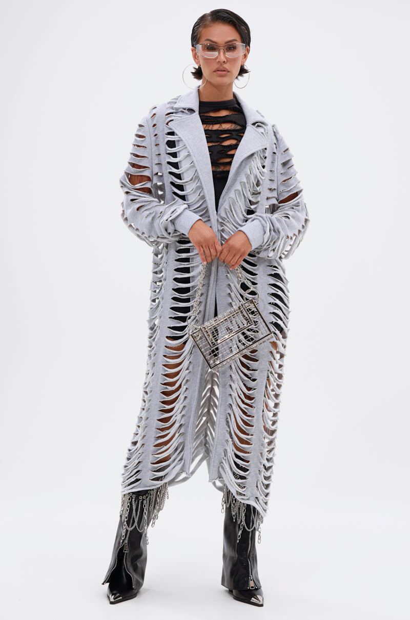 A person wears the CUT IT OUT FRENCH TERRY TRENCH in light gray over a black outfit, paired with black pointed boots featuring chain details, clear glasses, and carries a small rectangular handbag while posing against a white background. 3