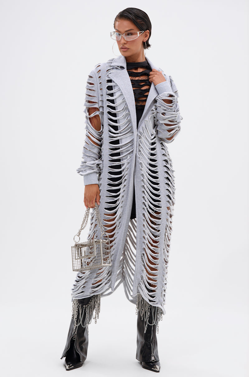 A woman wears the long, light gray CUT IT OUT FRENCH TERRY TRENCH over a black outfit, paired with clear glasses, a small metallic cage handbag, and shiny black boots against a plain white background. 4