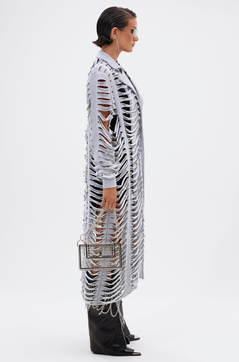 A person in profile wears the CUT IT OUT FRENCH TERRY TRENCH over black clothes, pointed black boots, and sunglasses, holding a geometric metallic handbag against a plain white background. 5