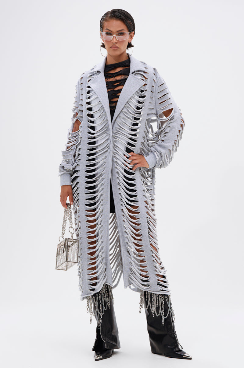 A woman wears clear sunglasses, the CUT IT OUT FRENCH TERRY TRENCH over a black outfit, black pointed boots, and carries a small silver handbag as she poses confidently against a plain white background. 6