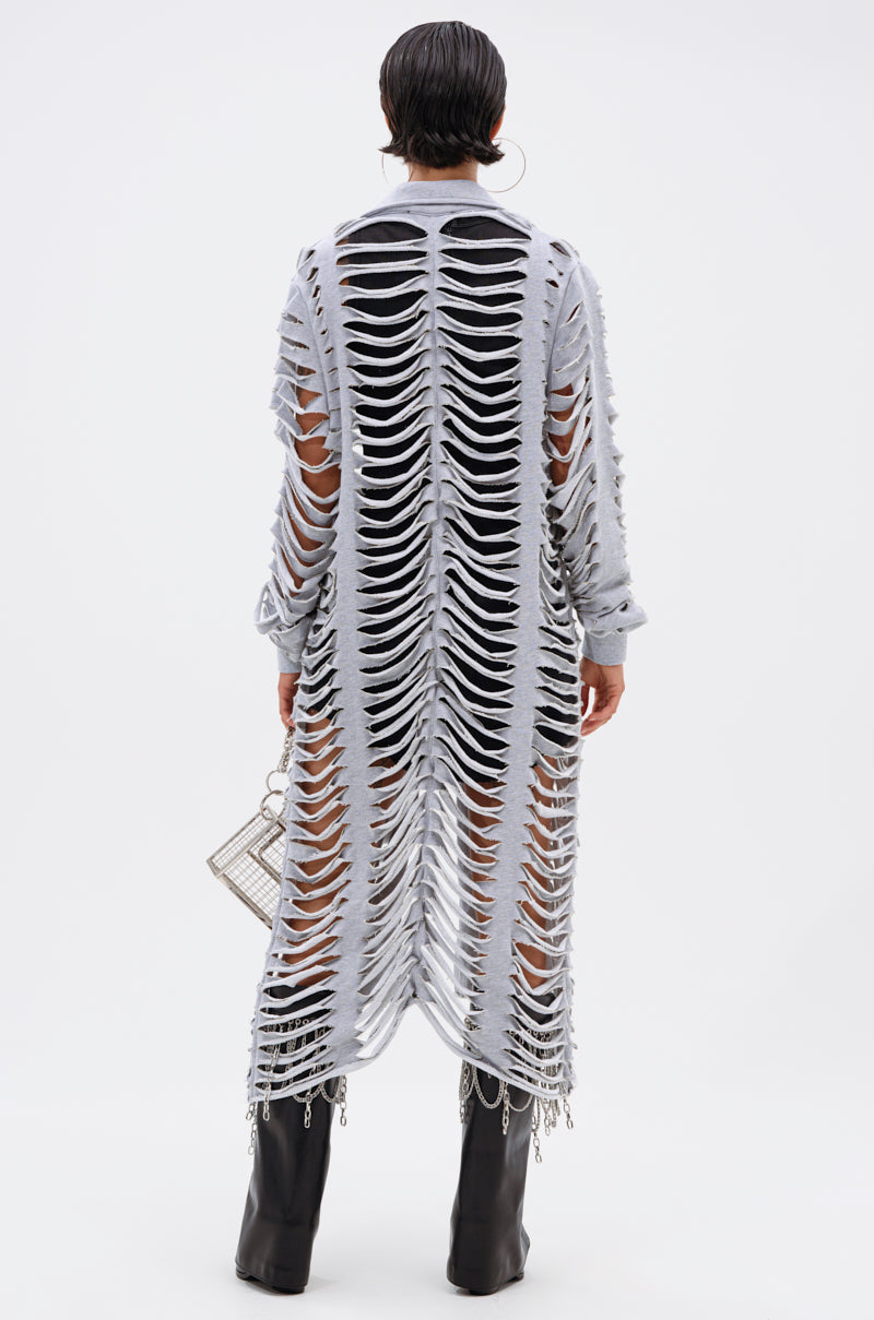 A person with short dark hair and large hoop earrings stands facing away in the CUT IT OUT FRENCH TERRY TRENCH—an edgy long gray knit coat with wide cutouts—styled with black boots and a small handbag for a bold look. 7