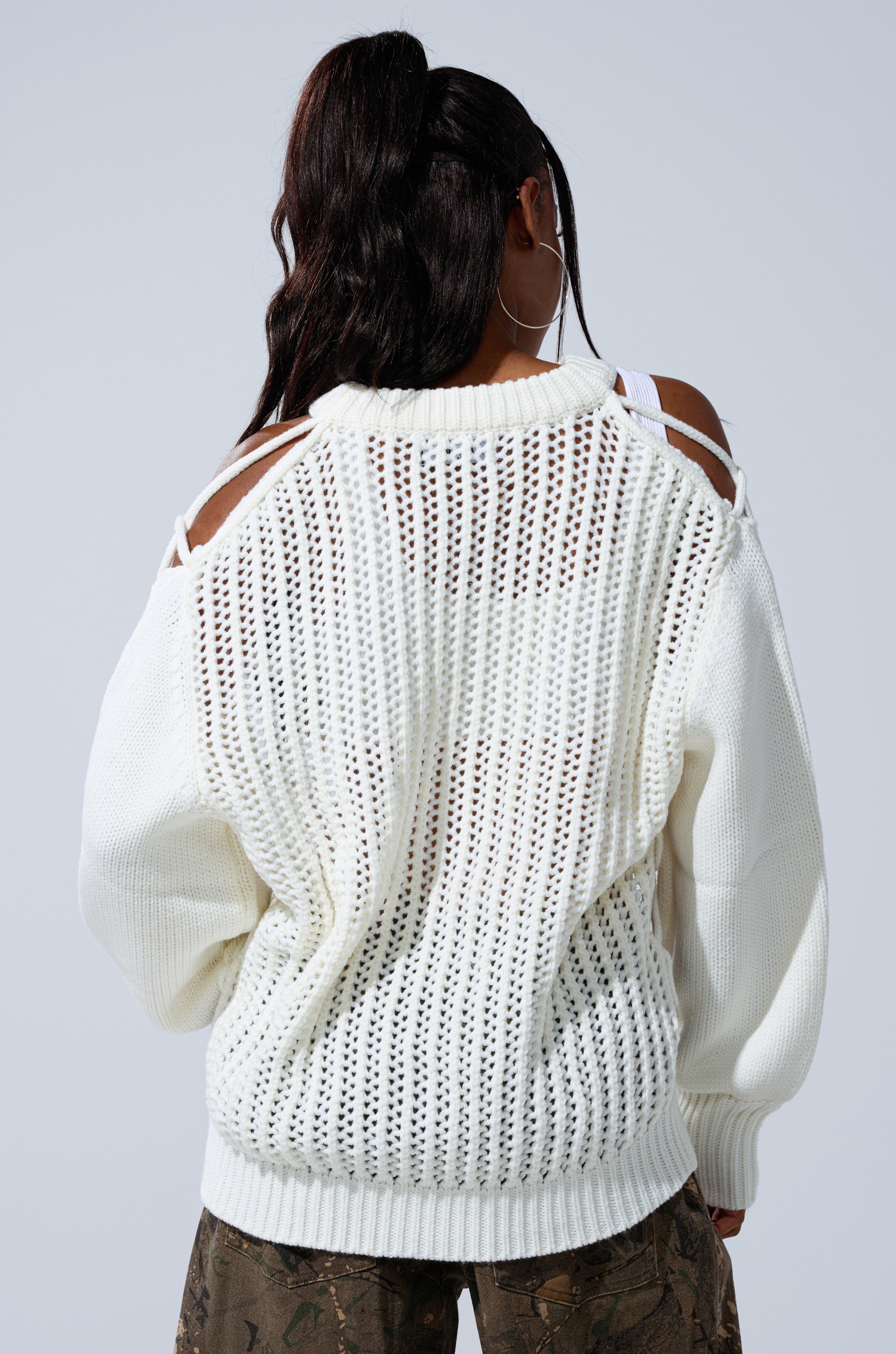 A person with long dark hair in a ponytail wears the OVER THE TOP CUT OUT OVERSIZED SWEATER and camouflage pants, shown from the back against a plain light background.