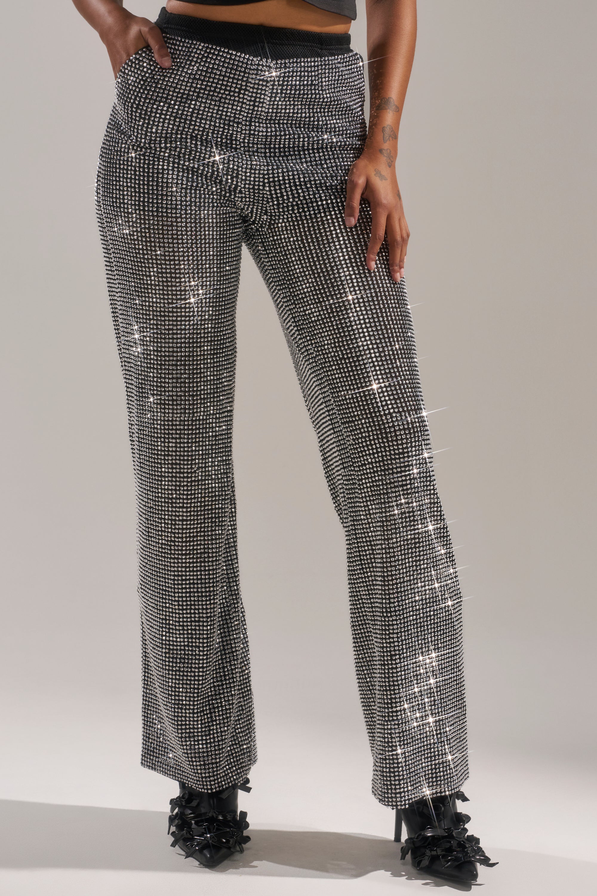 A person models the LET IT GO RHINESTONE WIDE LEG PANT, standing with one hand in their pocket and wearing black ruffled heels against a plain background.