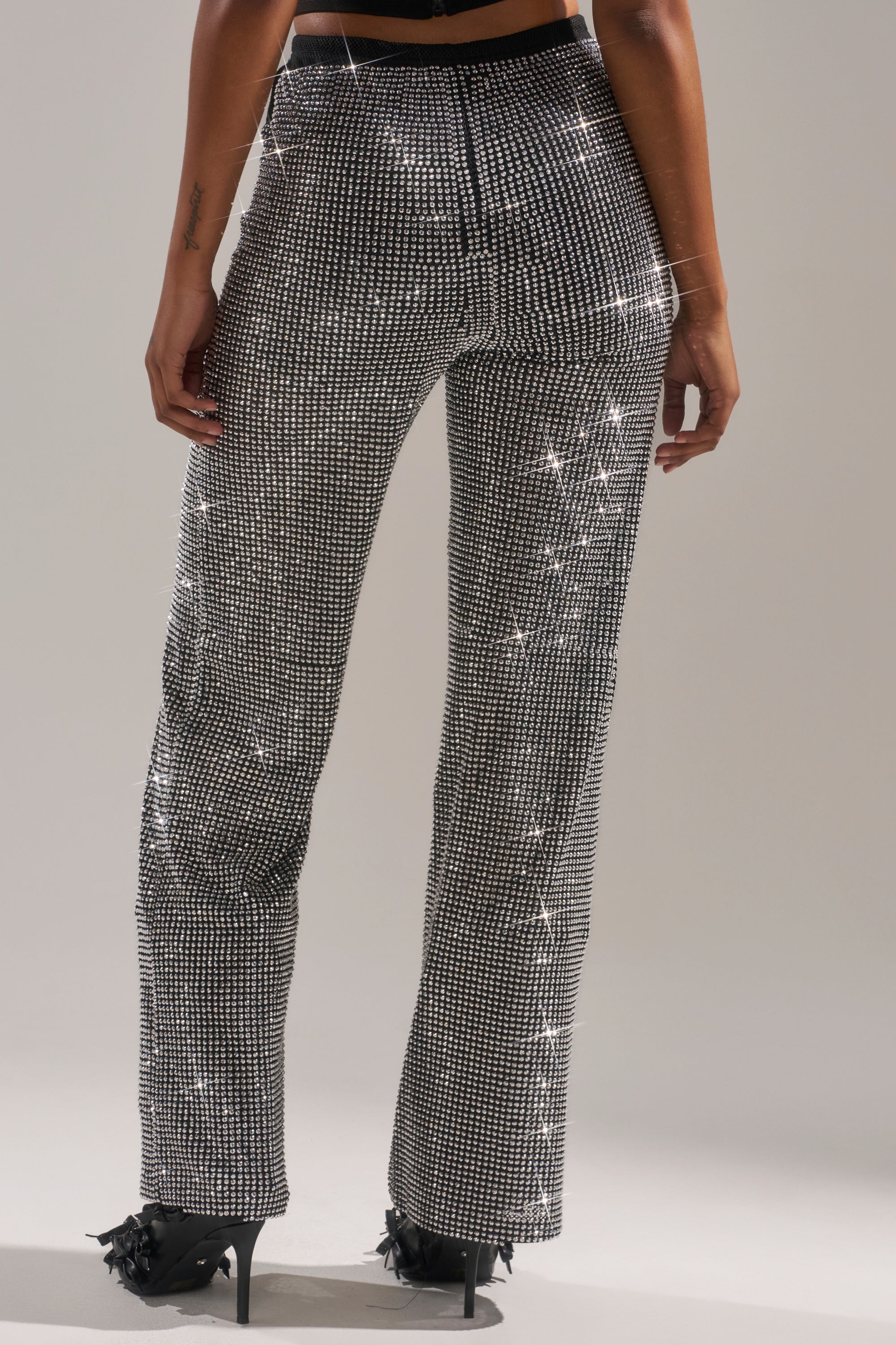 A person shown from the back wears the LET IT GO RHINESTONE WIDE LEG PANT—high-waisted and glittering with rhinestones—paired with black high heels against a neutral background.