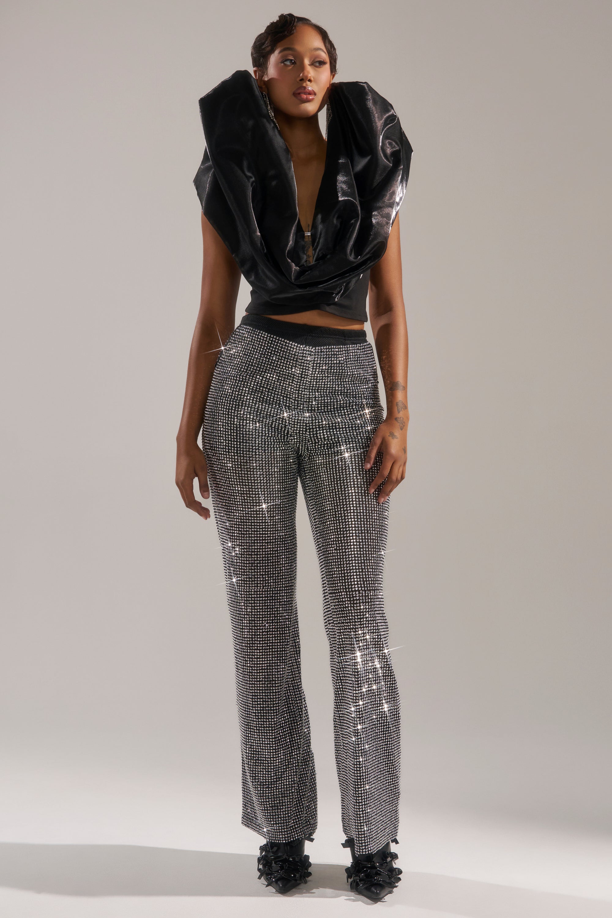 A woman stands confidently in the LET IT GO RHINESTONE WIDE LEG PANT, paired with a sculptural black top and textured black shoes, set against a plain light background.