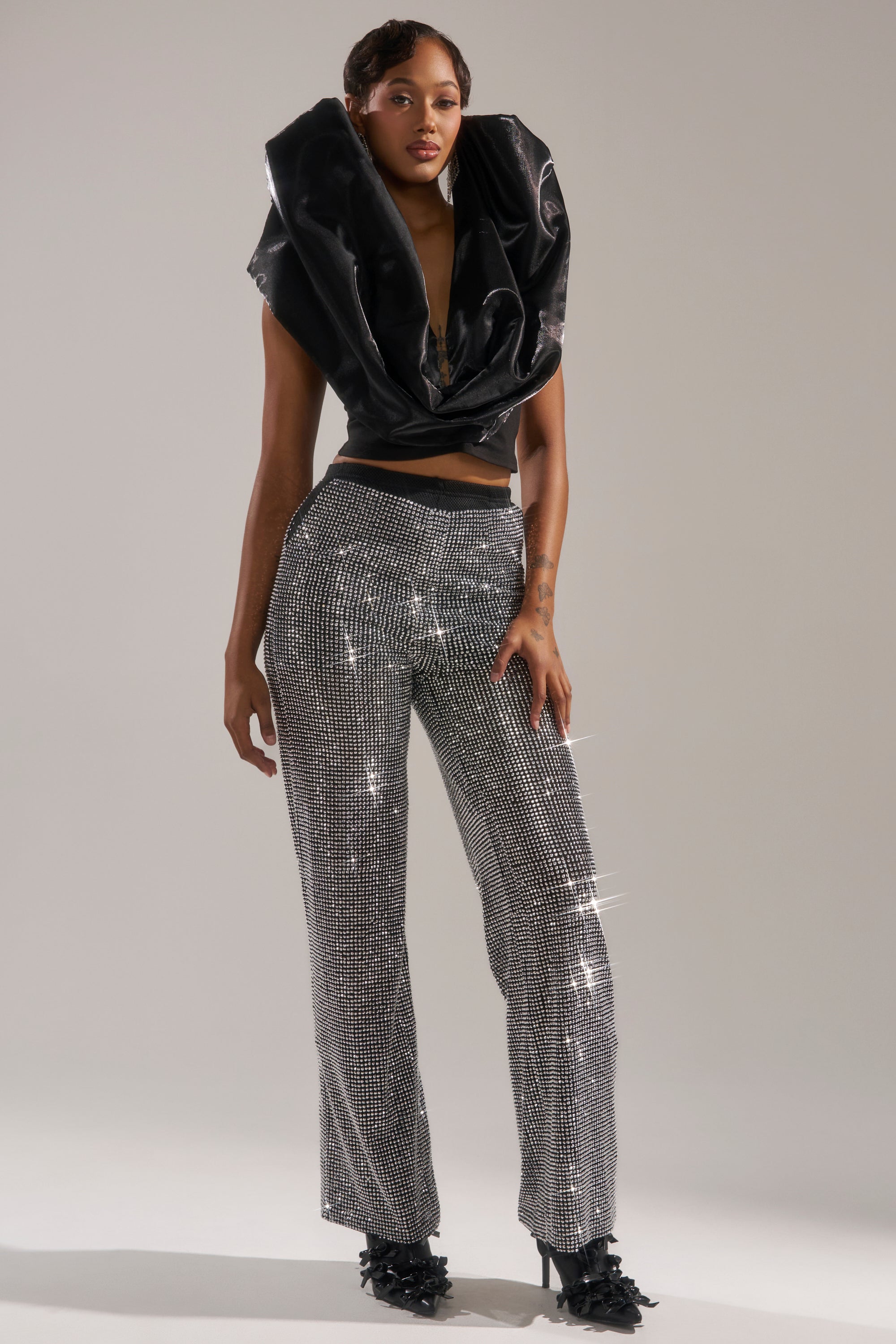 A person stands confidently in the LET IT GO RHINESTONE WIDE LEG PANT, paired with black feather-detailed high heels and a dramatic black sculptural top with a wide collar, against a softly lit plain background.