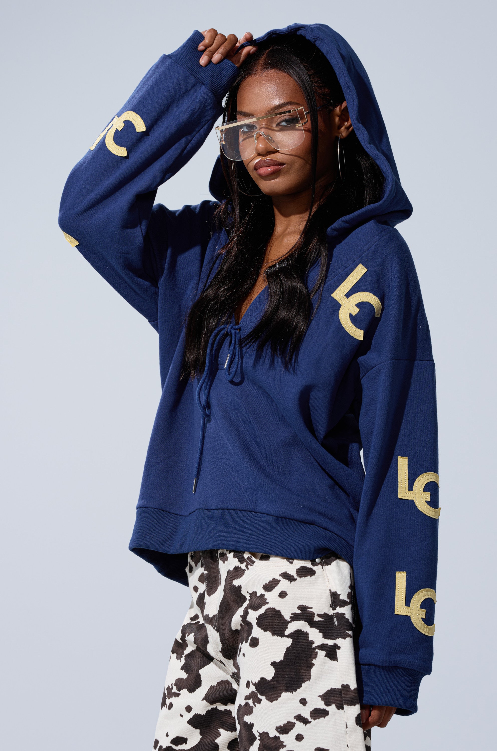 A woman models the CITY LOVER OVERSIZED SWEATSHIRT, a cozy cotton-blend piece with "LC" NYC Lovers Club logos. She pairs it with clear glasses and black-and-white cow print pants for a stylish look against a plain backdrop.