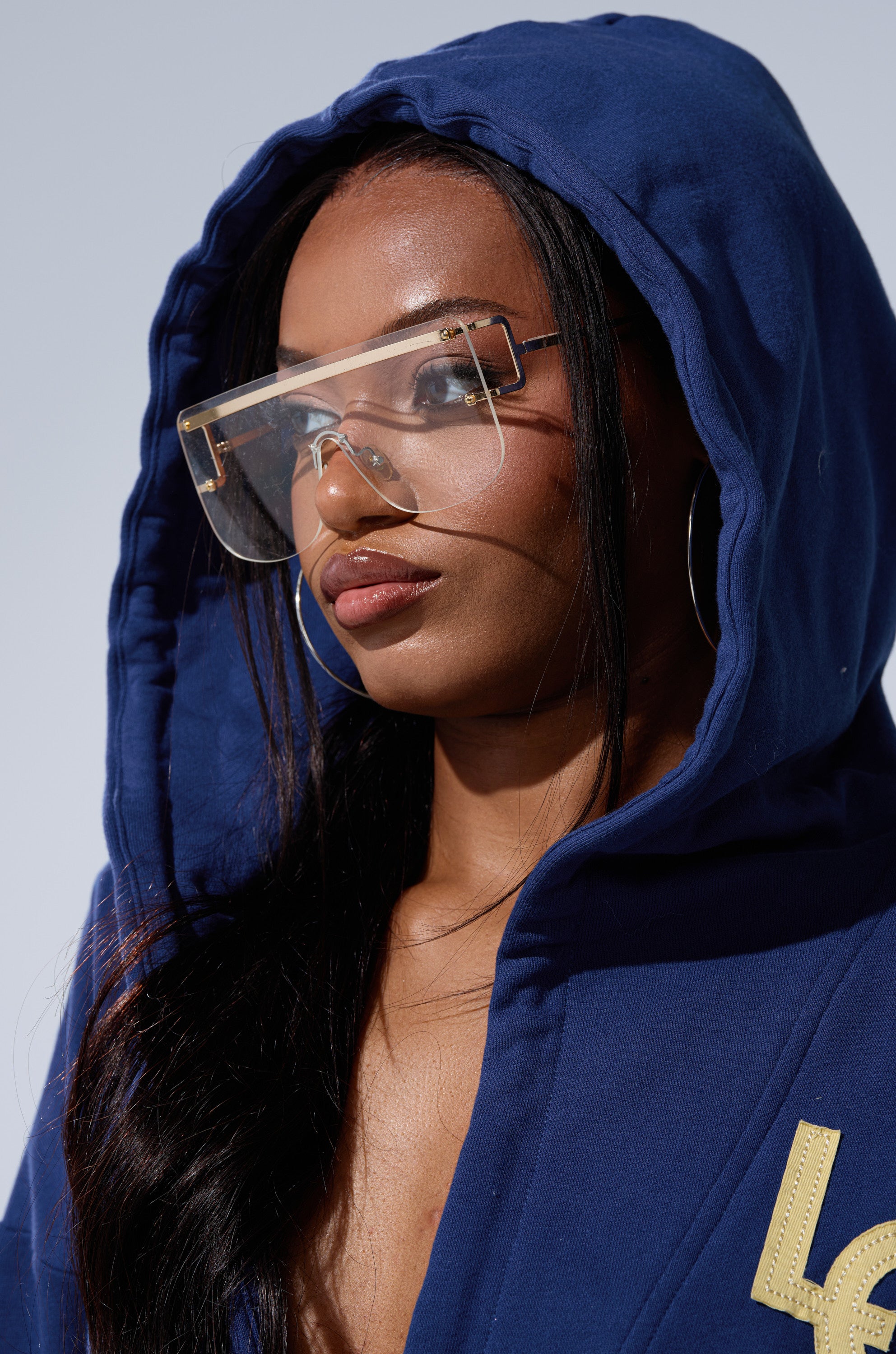 A woman with long dark hair and hoop earrings wears the CITY LOVER OVERSIZED SWEATSHIRT, a cozy blue cotton-blend hoodie, paired with large clear glasses and glossy lips. She looks ahead against a plain, light backdrop.