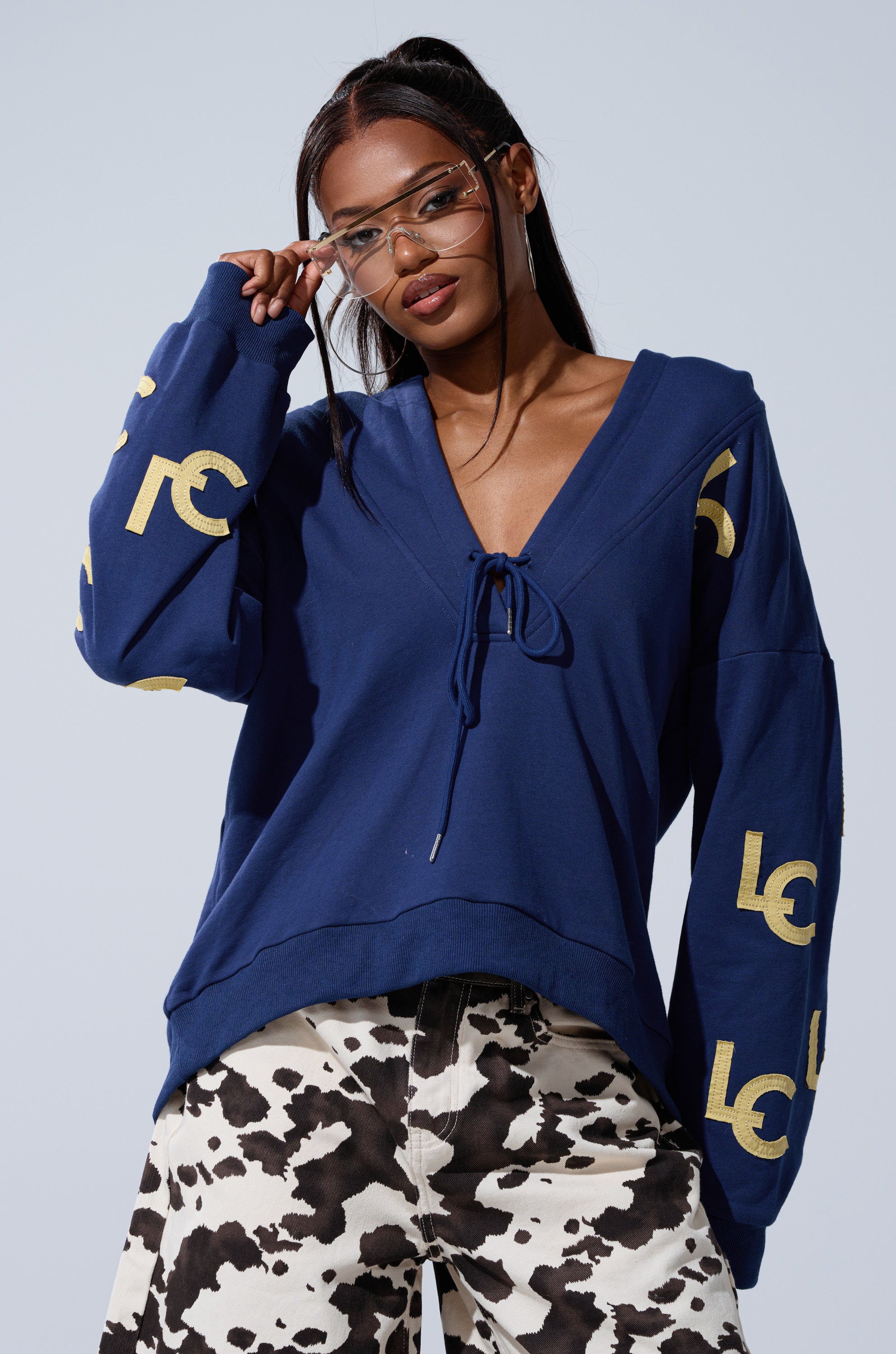 A woman poses confidently against a light background in the CITY LOVER OVERSIZED SWEATSHIRT—an oversized, cozy blue cotton-blend with gold sleeve letters—paired with clear glasses and cow print pants; perfect for NYC lovers.