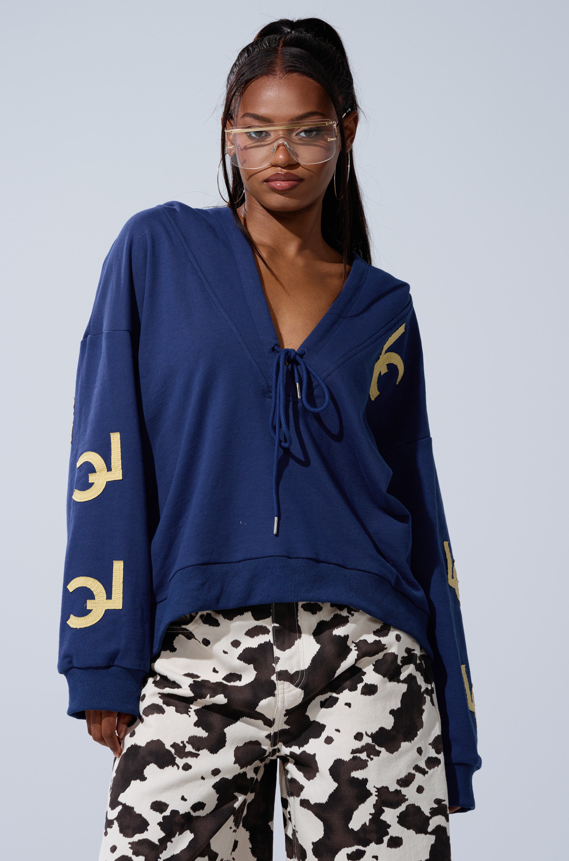 A woman in clear glasses and black-and-white cow print pants stands against a light gray background, wearing the CITY LOVER OVERSIZED SWEATSHIRT—a blue hoodie featuring yellow sleeve designs from the NYC Lovers Club.