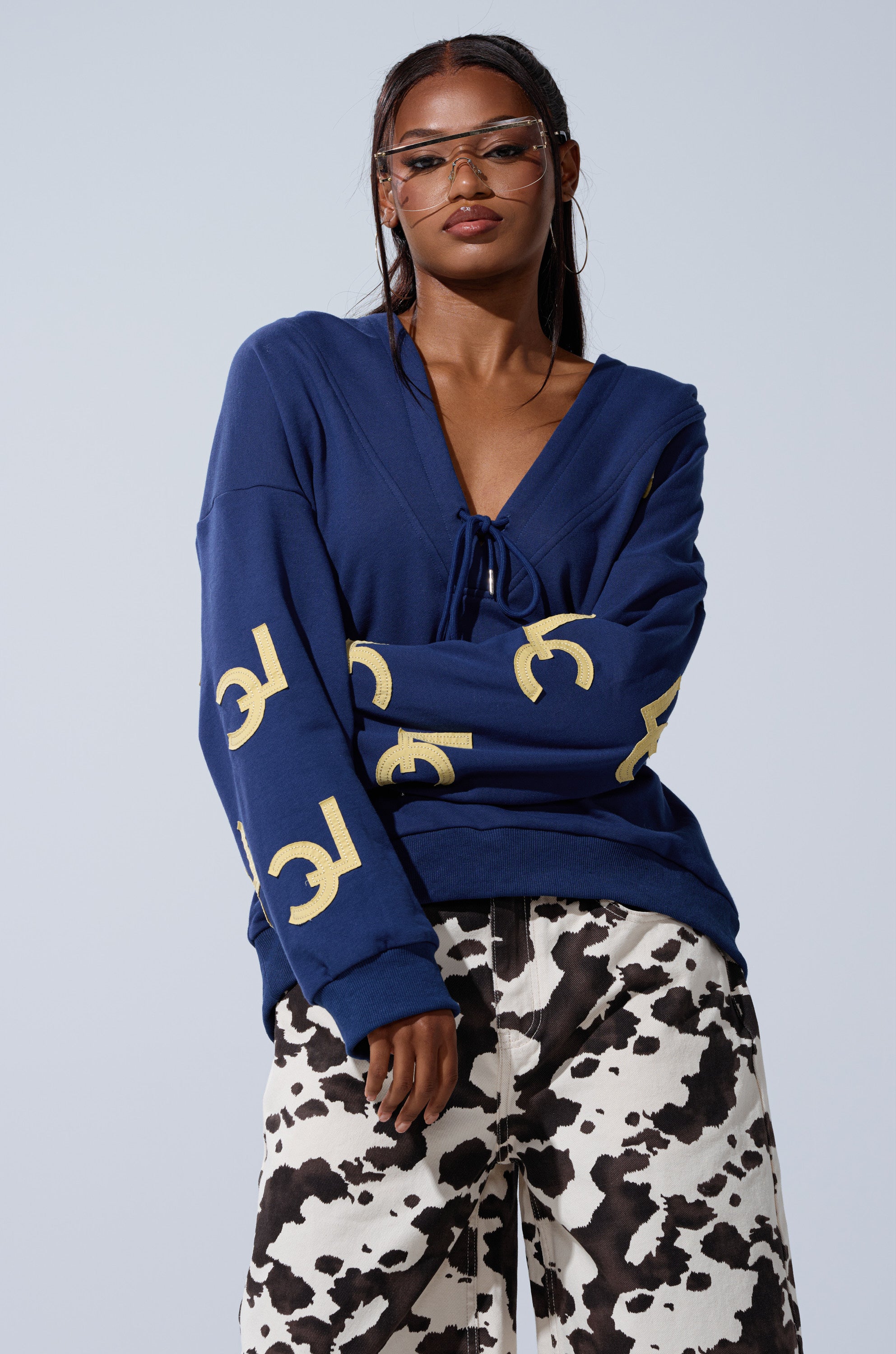 A woman in clear glasses wears the CITY LOVER OVERSIZED SWEATSHIRT with yellow sleeve designs and black-and-white cow print pants, posing against a plain light background.