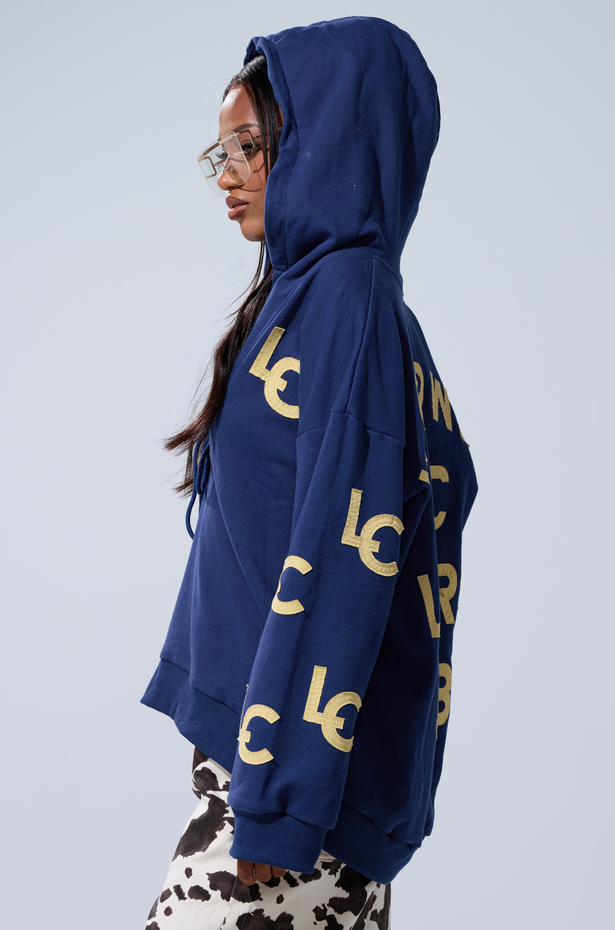 A woman in a blue CITY LOVER OVERSIZED SWEATSHIRT with yellow "LC" lettering, cow-print pants, and clear glasses stands in profile against a plain background—ideal for NYC Lovers Club fans embracing bold fashion.