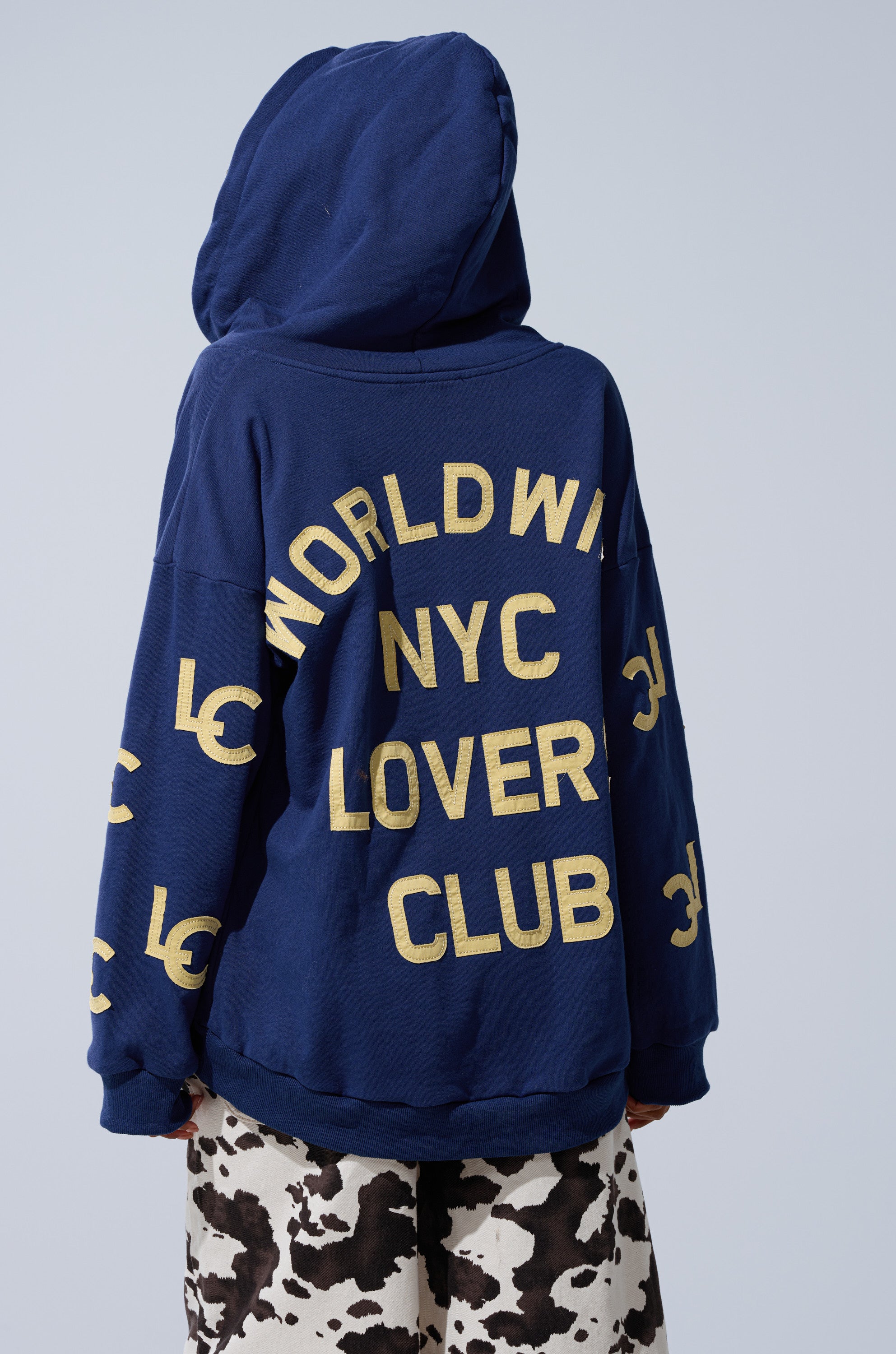 Someone wears the CITY LOVER OVERSIZED SWEATSHIRT, a cozy cotton-blend hoodie with bold yellow "WORLDWIDE NYC LOVERS CLUB" text on the back, hood up, paired with black and white patterned pants.