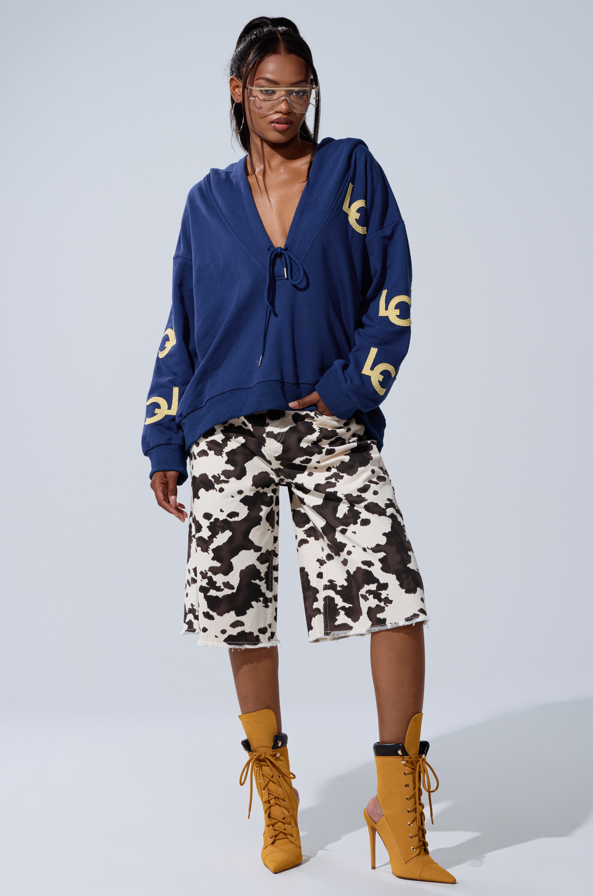 A woman models the CITY LOVER OVERSIZED SWEATSHIRT with yellow designs, cow-print knee-length shorts, mustard lace-up heeled boots, and clear glasses—ideal for NYC club style.