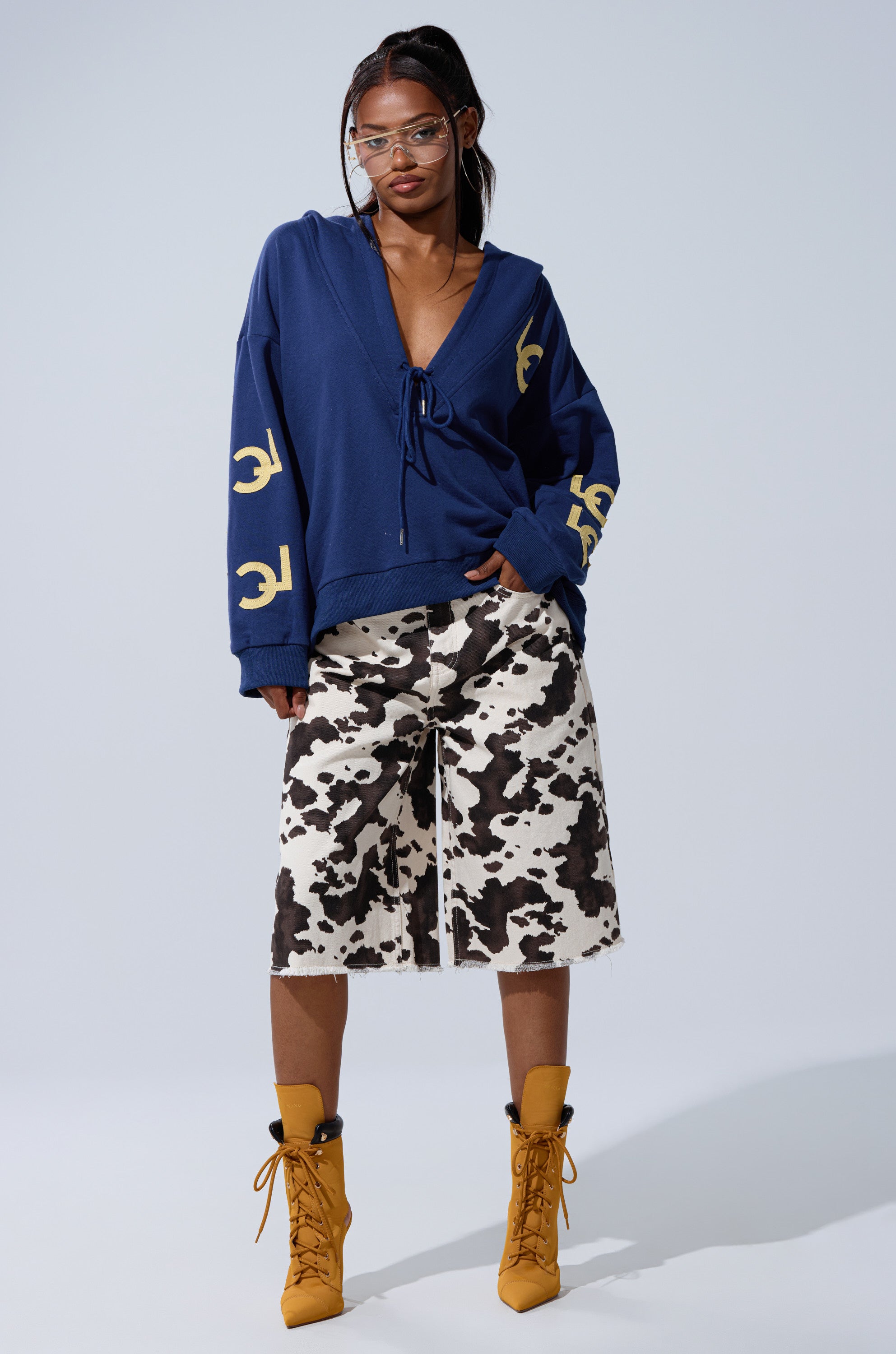 A woman stands confidently in the CITY LOVER OVERSIZED SWEATSHIRT—blue, cozy, and cotton-blend with yellow sleeve designs—paired with black-and-white cow print knee-length shorts, mustard-yellow lace-up boots, and clear glasses.