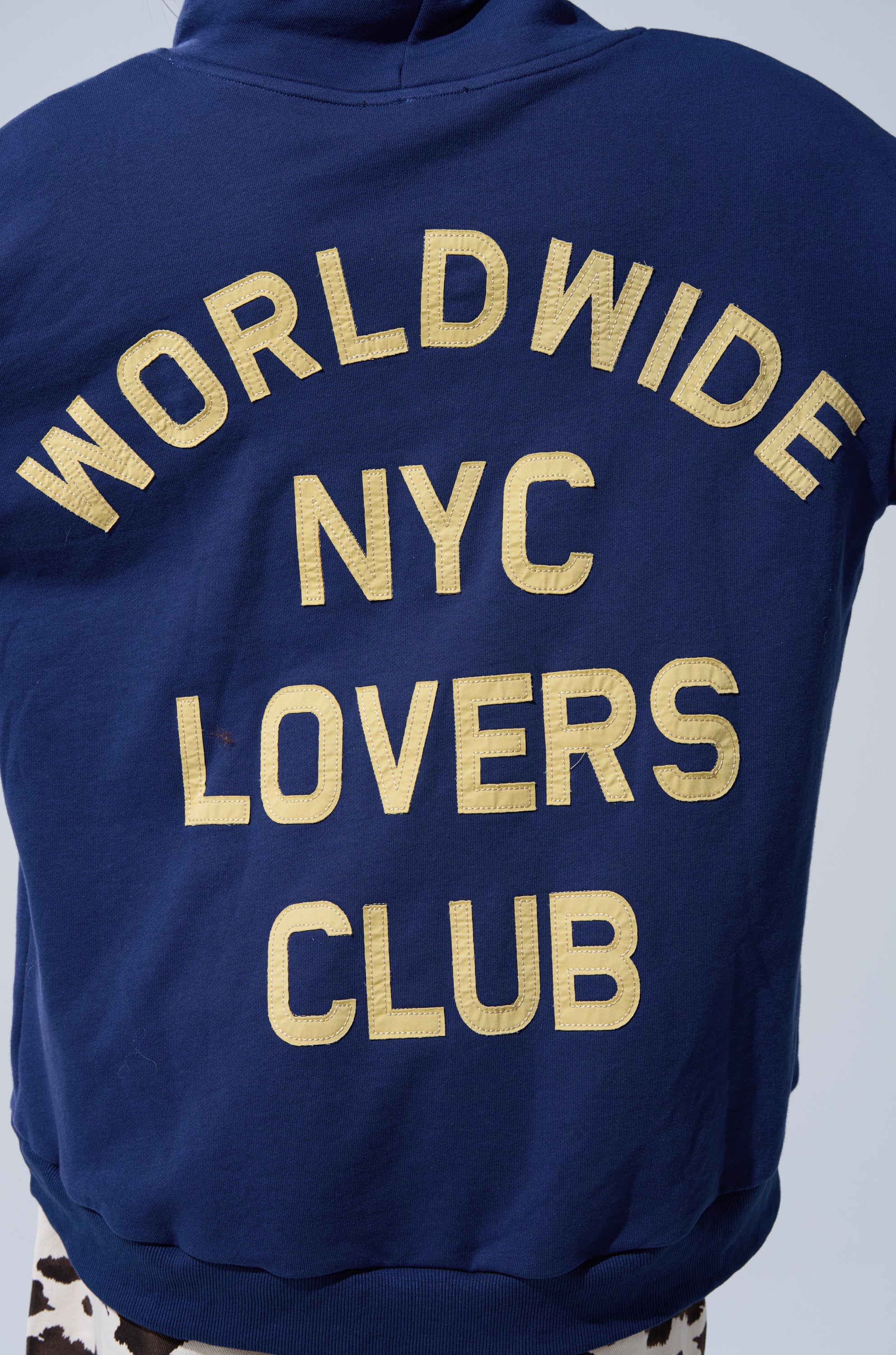 A person wearing the CITY LOVER OVERSIZED SWEATSHIRT, a blue cotton-blend with bold yellow "WORLDWIDE NYC LOVERS CLUB" lettering on the back, is turned away to show only their back.
