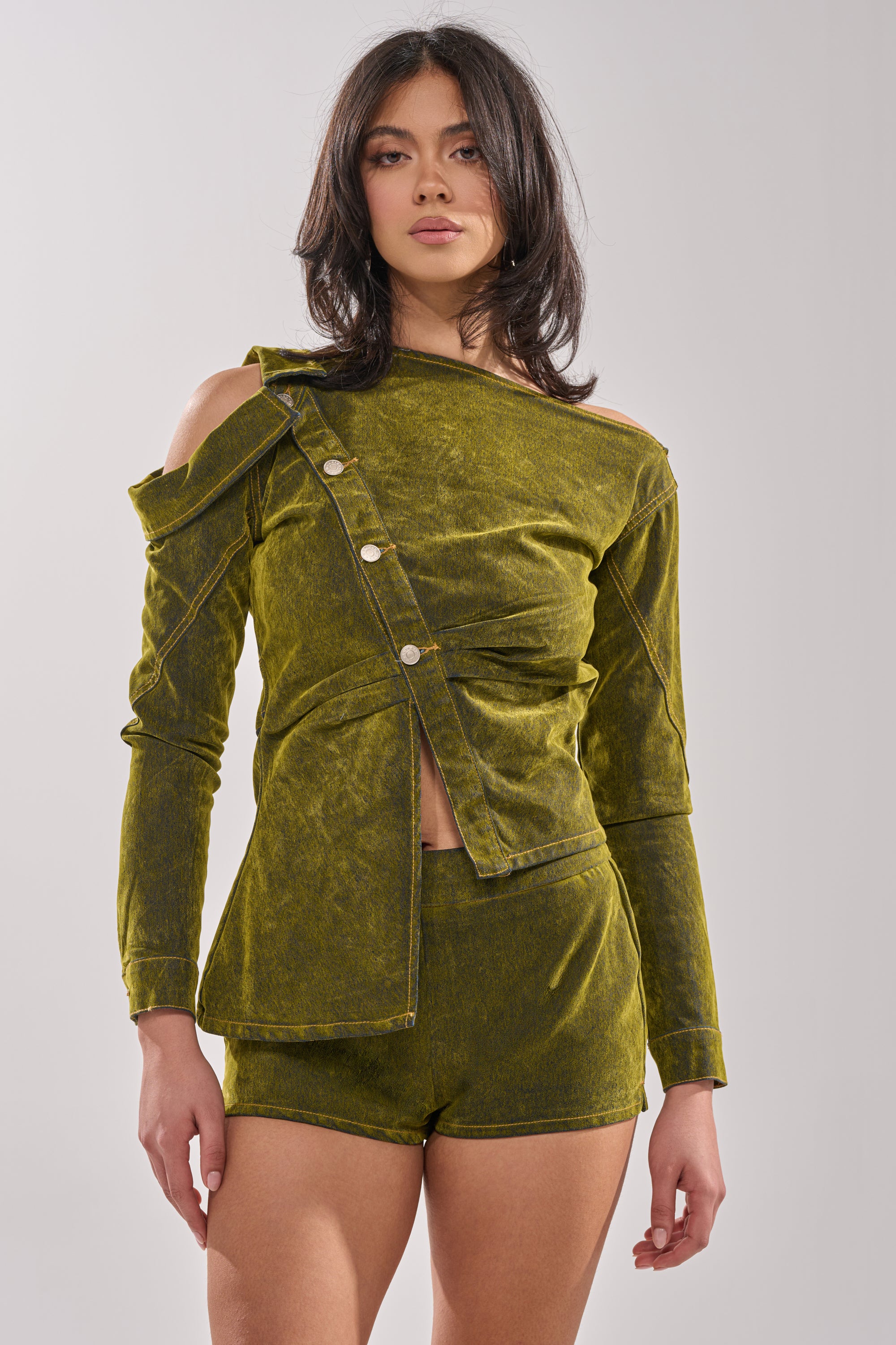 A woman with medium-length dark hair models the YOUR TIME IS COMING VELVET DENIM BLOUSE—an olive green, asymmetrical, off-shoulder long-sleeve top with buttons—paired with matching high-waisted shorts against a plain light background. 0