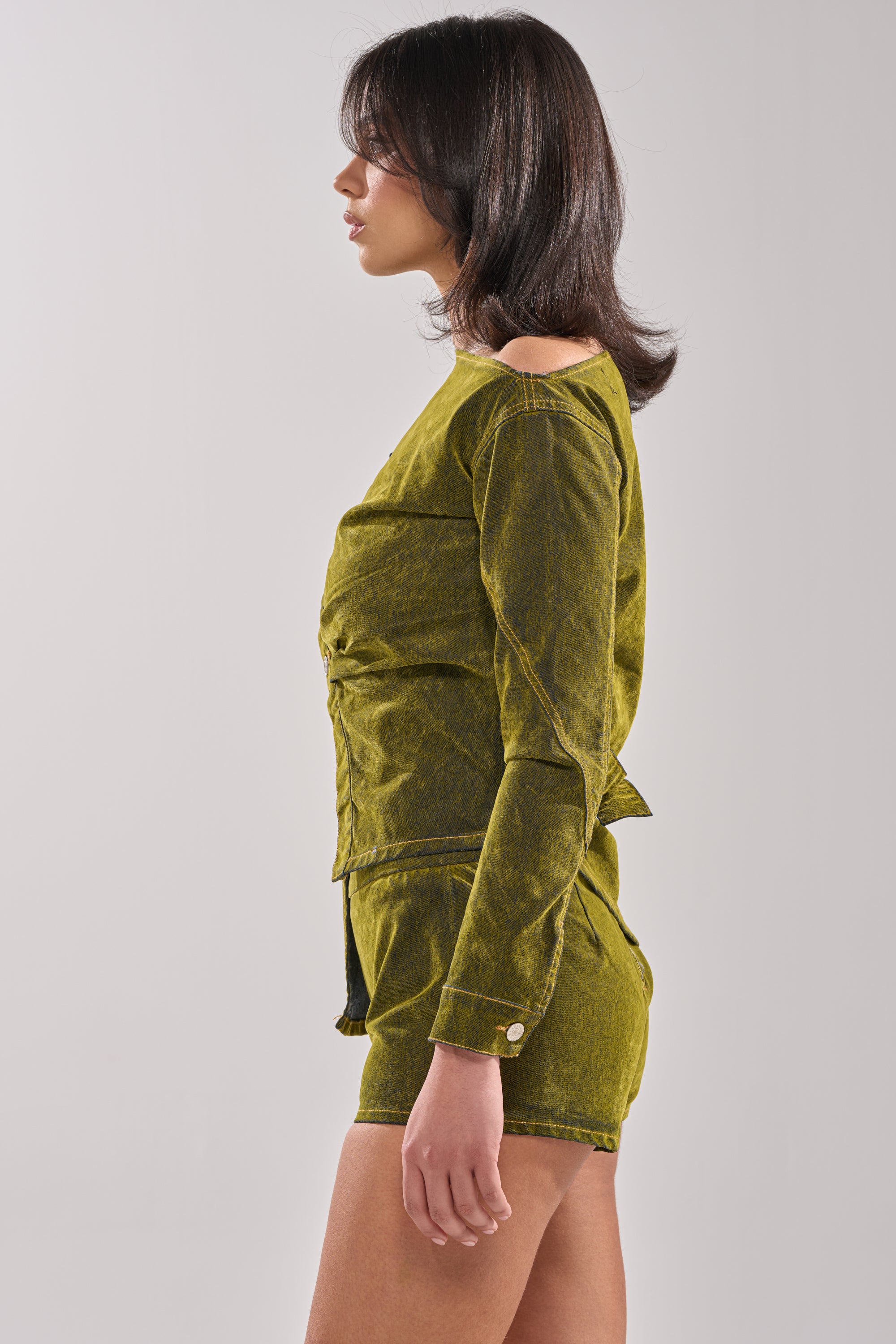 A woman with shoulder-length dark hair stands in profile, wearing the YOUR TIME IS COMING VELVET DENIM BLOUSE, a sculpted olive green long-sleeve top, set against a plain light gray background. 3