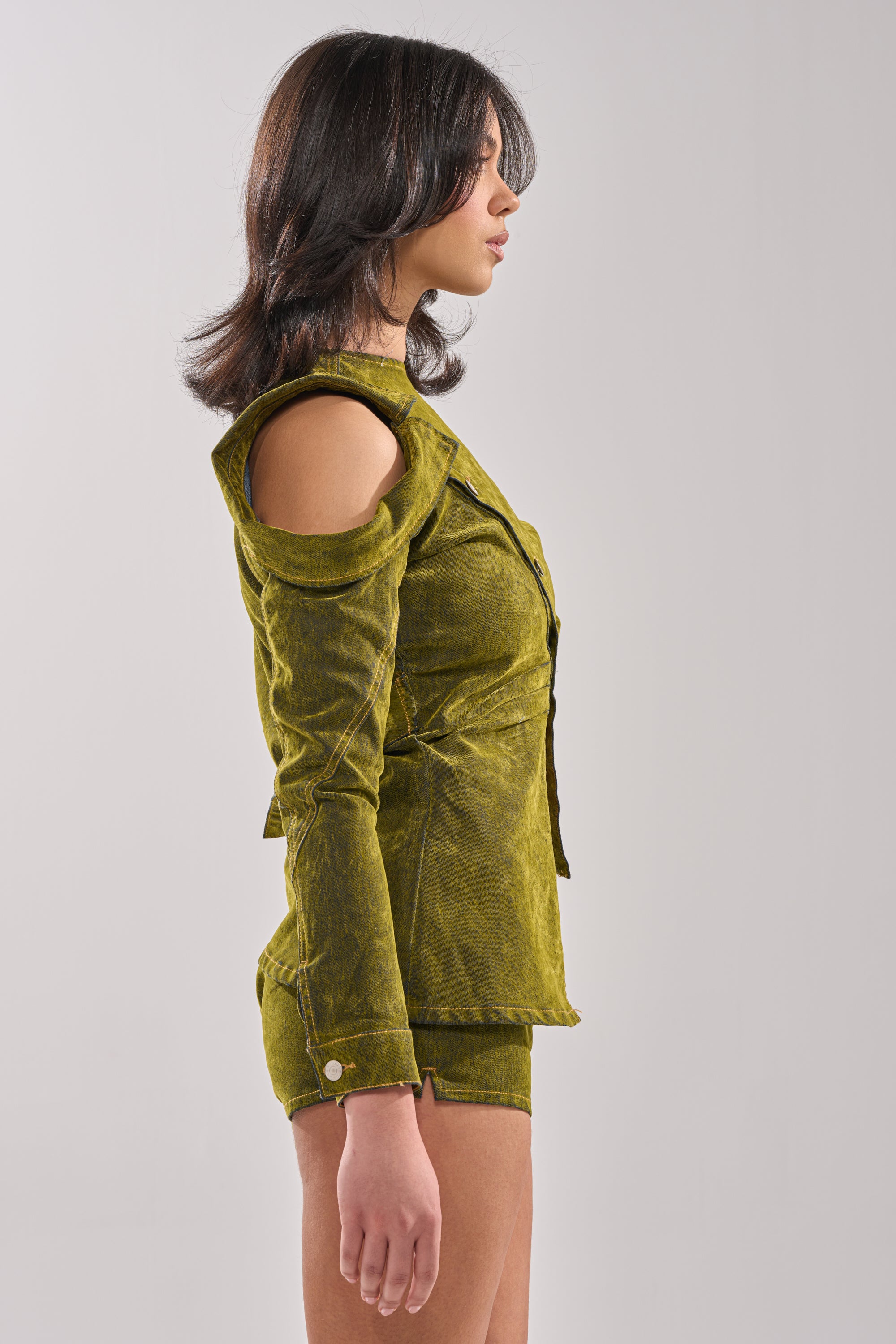A woman in profile wears the YOUR TIME IS COMING VELVET DENIM BLOUSE, featuring mustard green velvet, an asymmetrical neckline, and cold shoulder sleeves, set against a plain light gray background. 4