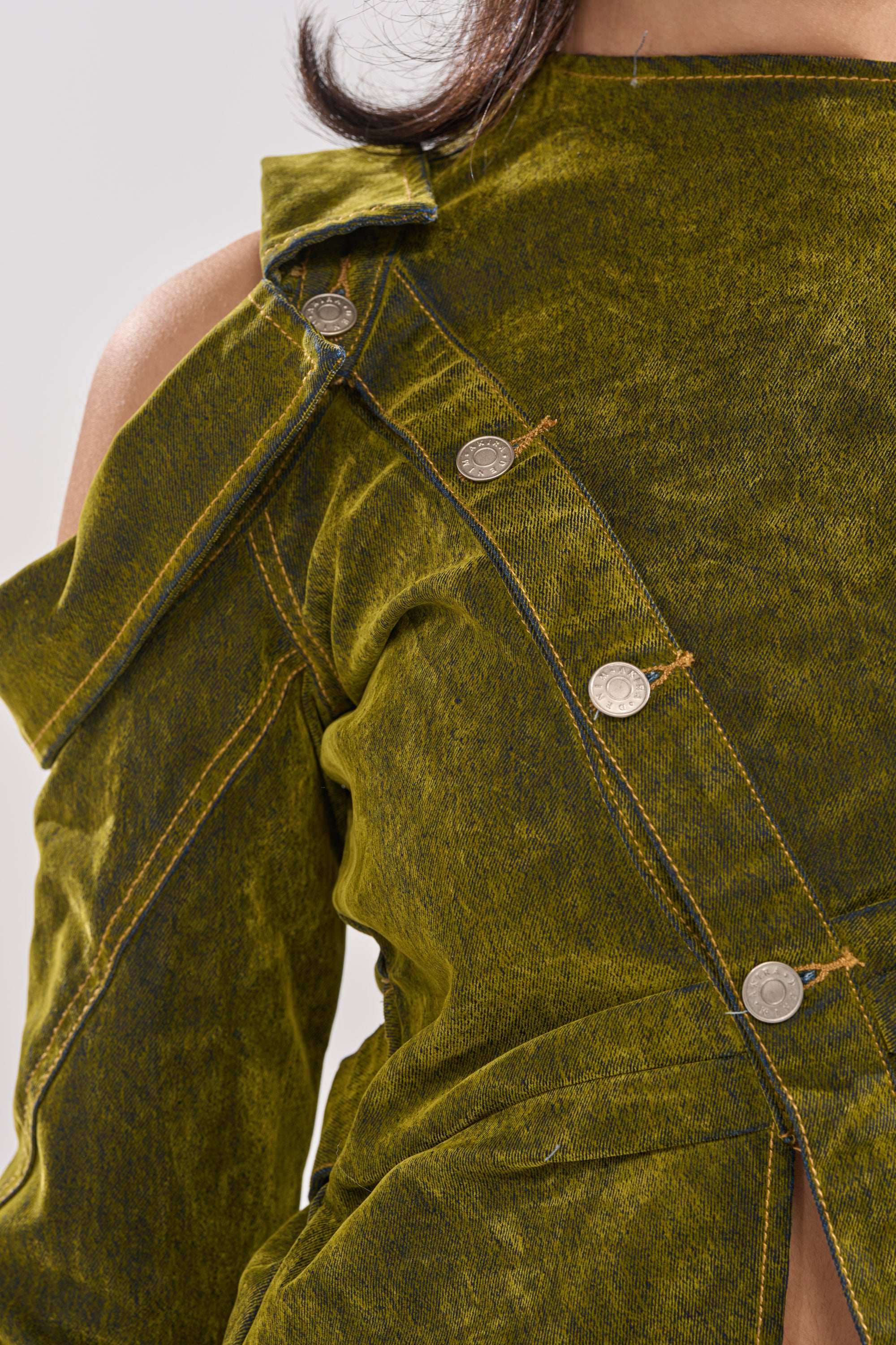 Close-up of a person wearing the "YOUR TIME IS COMING VELVET DENIM BLOUSE" in olive green, featuring textured fabric, an asymmetrical neckline, diagonal metallic buttons, visible seams, and a sculpted modern fit. 7