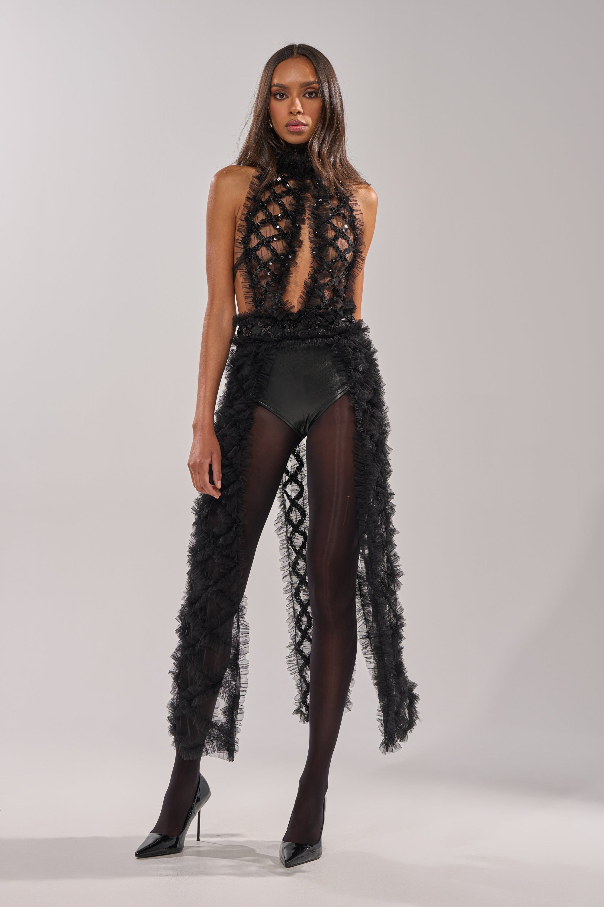 A woman exudes confidence in the BRING ON THE DRAMA BLOUSE—an eye-catching black, textured top with sheer panels and fringed accents, styled with black tights and high heels against a plain gray background. 0