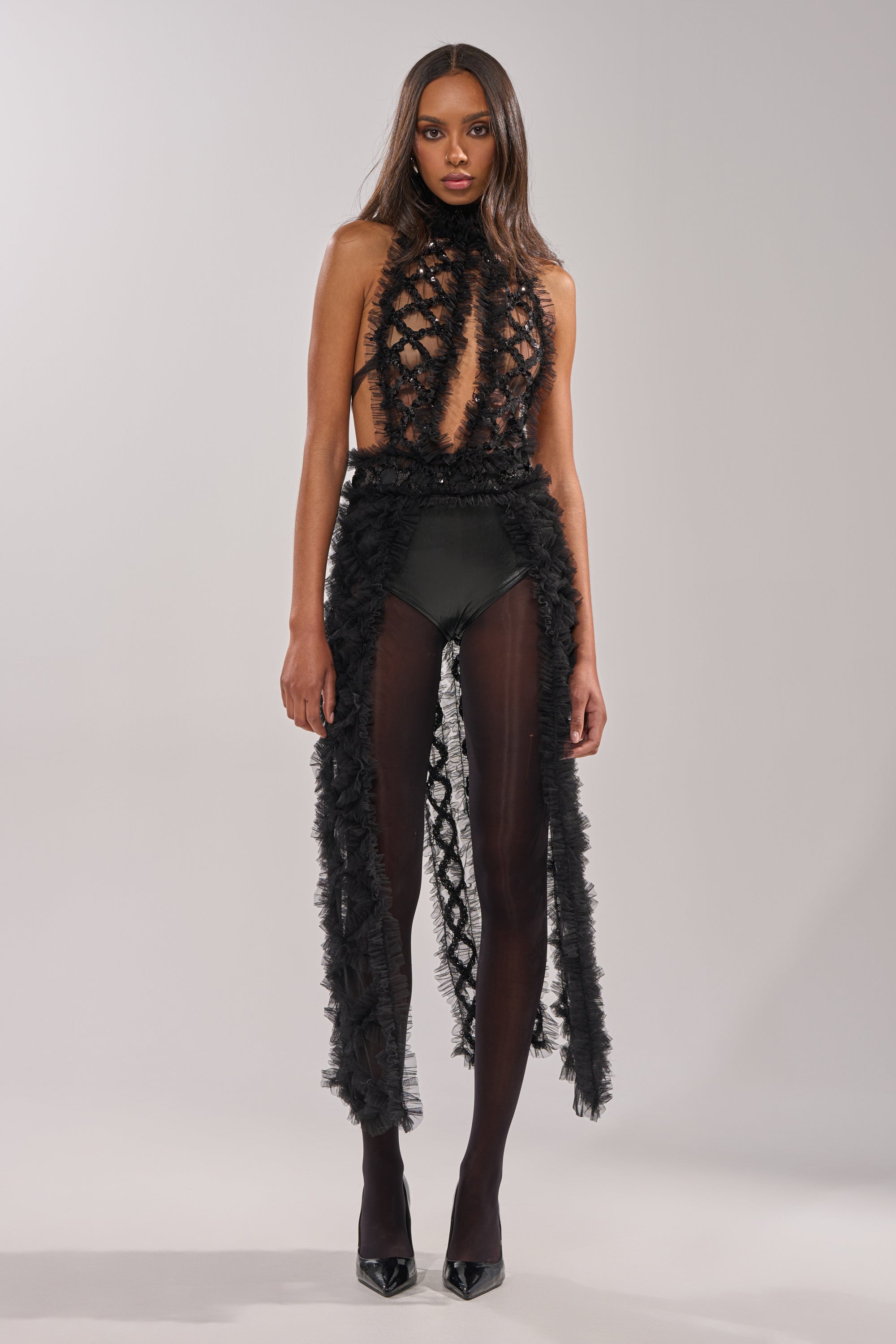 A woman faces forward in the BRING ON THE DRAMA BLOUSE, a bold black festival top featuring a sheer netted bodice and dramatic tulle accents. She has long straight hair, high-waisted bottoms, and wears black pointed shoes. 3