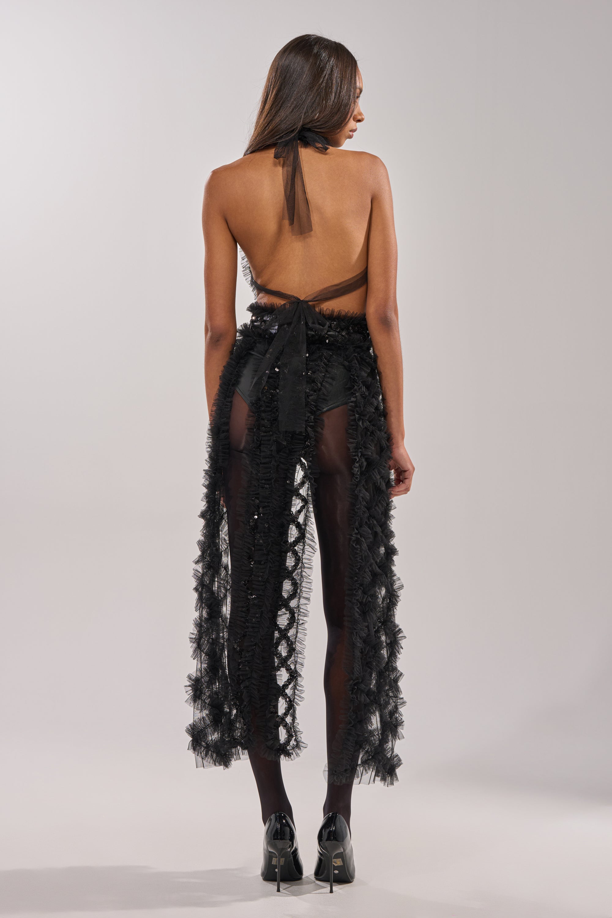 A woman with long hair faces away, wearing the BRING ON THE DRAMA BLOUSE paired with a sheer black ruffled skirt and black high heels, set against a neutral background. 6