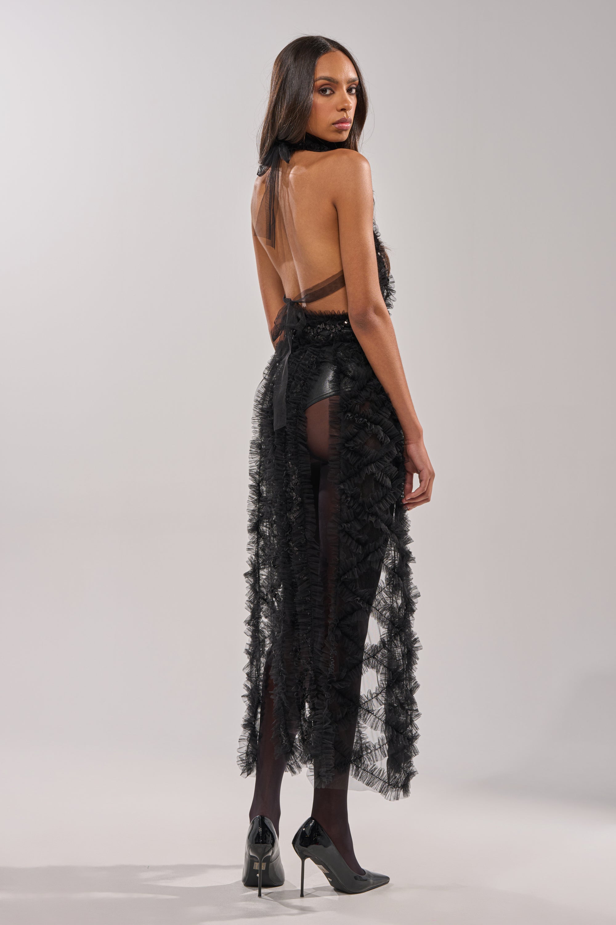 A woman with long, straight hair stands facing backward, looking over her shoulder. She wears the BRING ON THE DRAMA BLOUSE—a sheer sequin top—styled with black tights and heels against a neutral background. 7