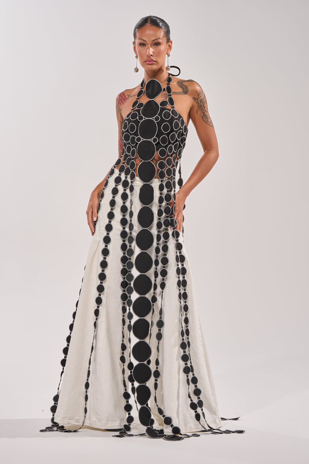 A woman models the DISILLUSIONED HALTER TOP, a black and white halter-neck design with cutout details of large black circles connected by smaller ones in vertical rows, standing hands on hips against a plain background. 0