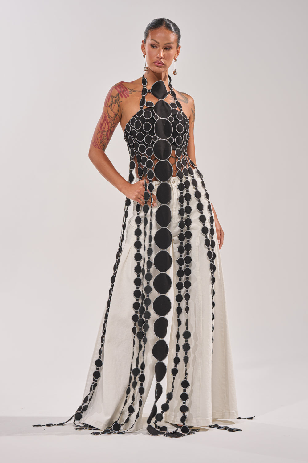 A woman models the DISILLUSIONED HALTER TOP, a sleeveless black and white gown featuring bold vertical circles, an open bodice with cutout detailing, and a long flowing skirt accented by striking circular motifs. 1