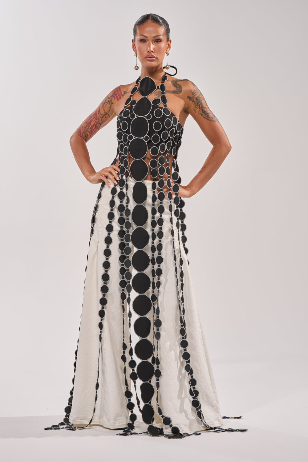 A woman poses confidently in the DISILLUSIONED HALTER TOP, featuring cutout details and a pattern of bold black circles on white. She wears statement earrings, displays arm tattoos, and stands against a plain light backdrop. 2