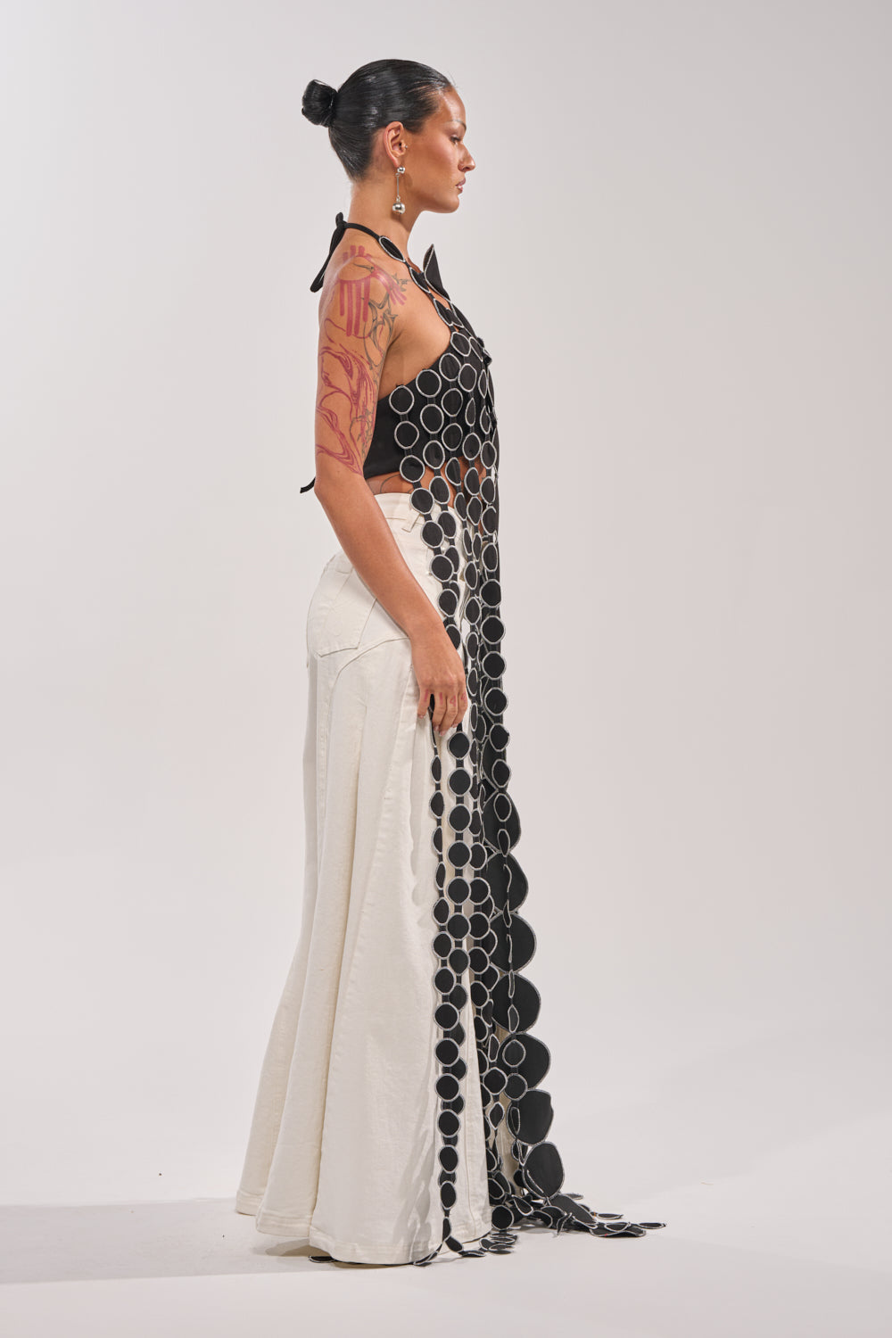 A person stands in profile against a plain background, wearing the black DISILLUSIONED HALTER TOP with circular black and silver embellishments and cutout details, paired with a long, high-waisted white skirt. Their dark hair is tied back in a bun. 3