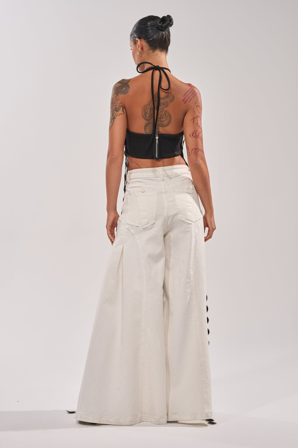 A tattooed person wears the DISILLUSIONED HALTER TOP, styled with wide-legged white pants. Their hair is tied up and the background is plain and light-colored. 4