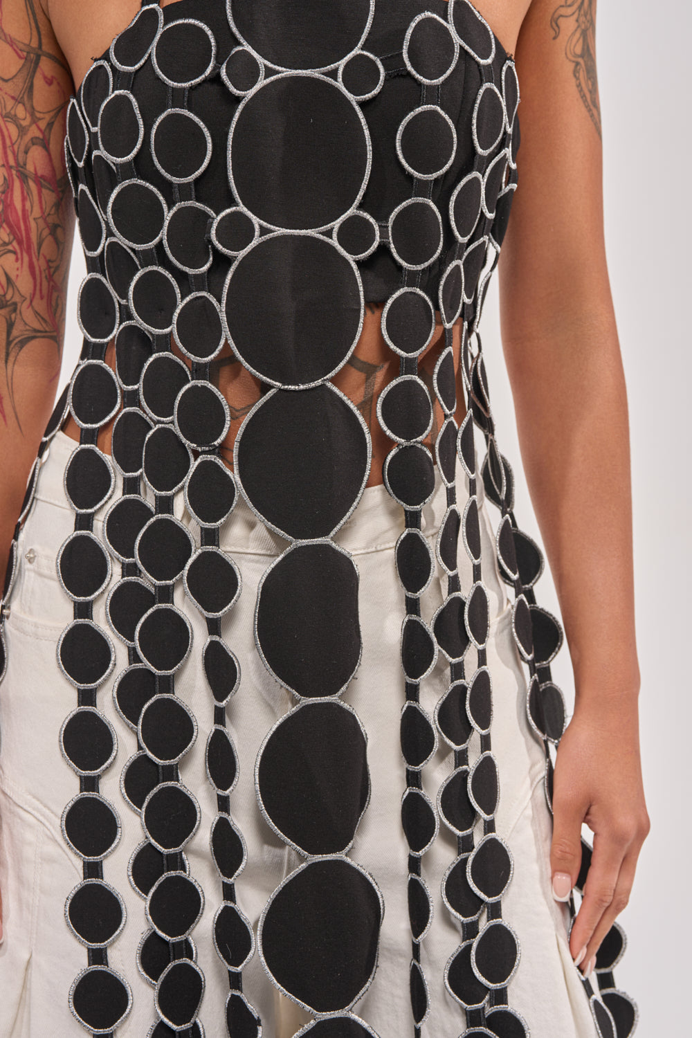 A person wears the DISILLUSIONED HALTER TOP, a sleeveless openwork appliqué piece with black and white circular patterns over light pants, showing tattooed arms. The top features a unique see-through circular design. 6