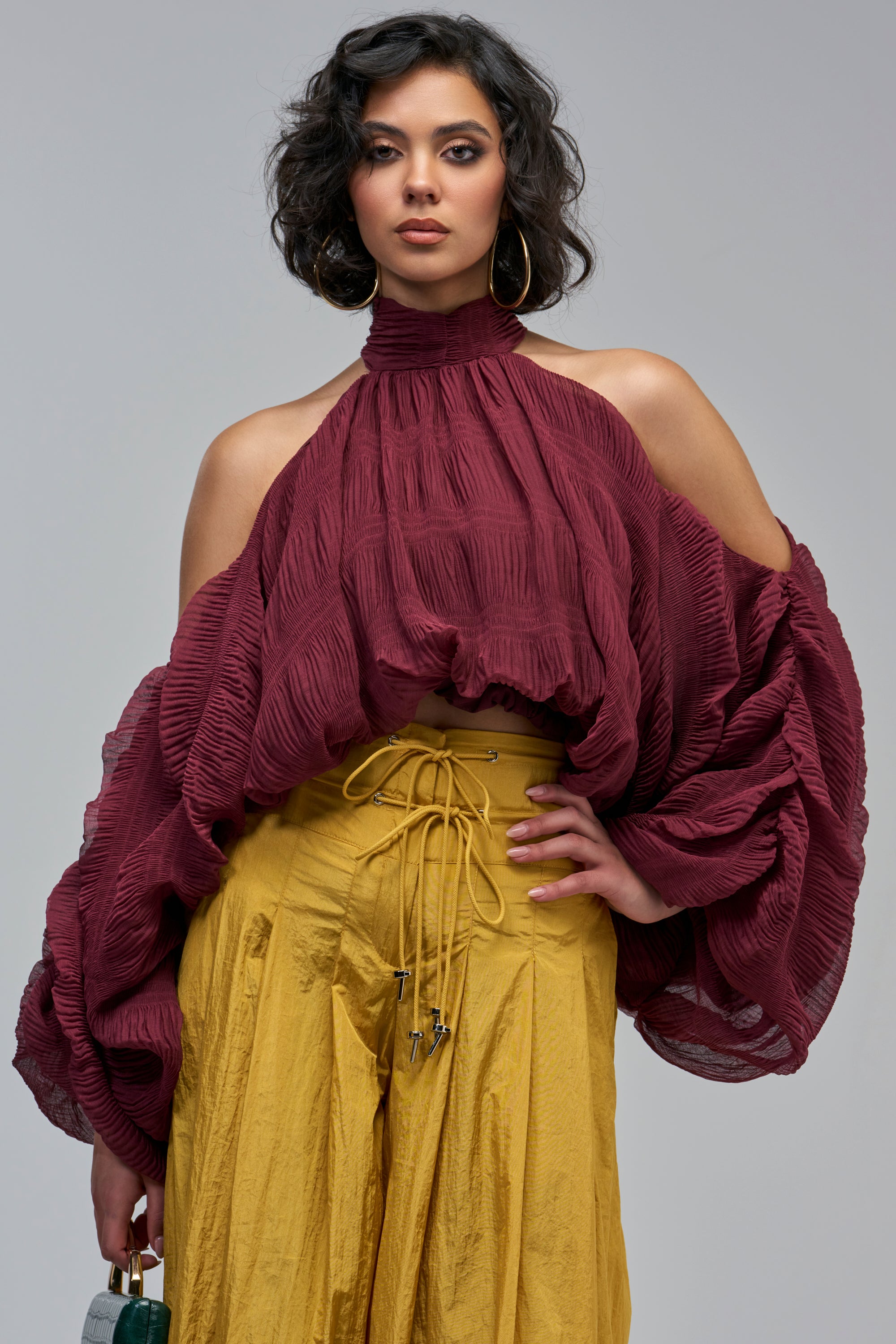 A model wears a SOFT DRAMA CHIFFON BLOUSE in burgundy, featuring a pleated, cold-shoulder design with a high neck and wide sleeves, paired with mustard yellow lace-up trousers.
