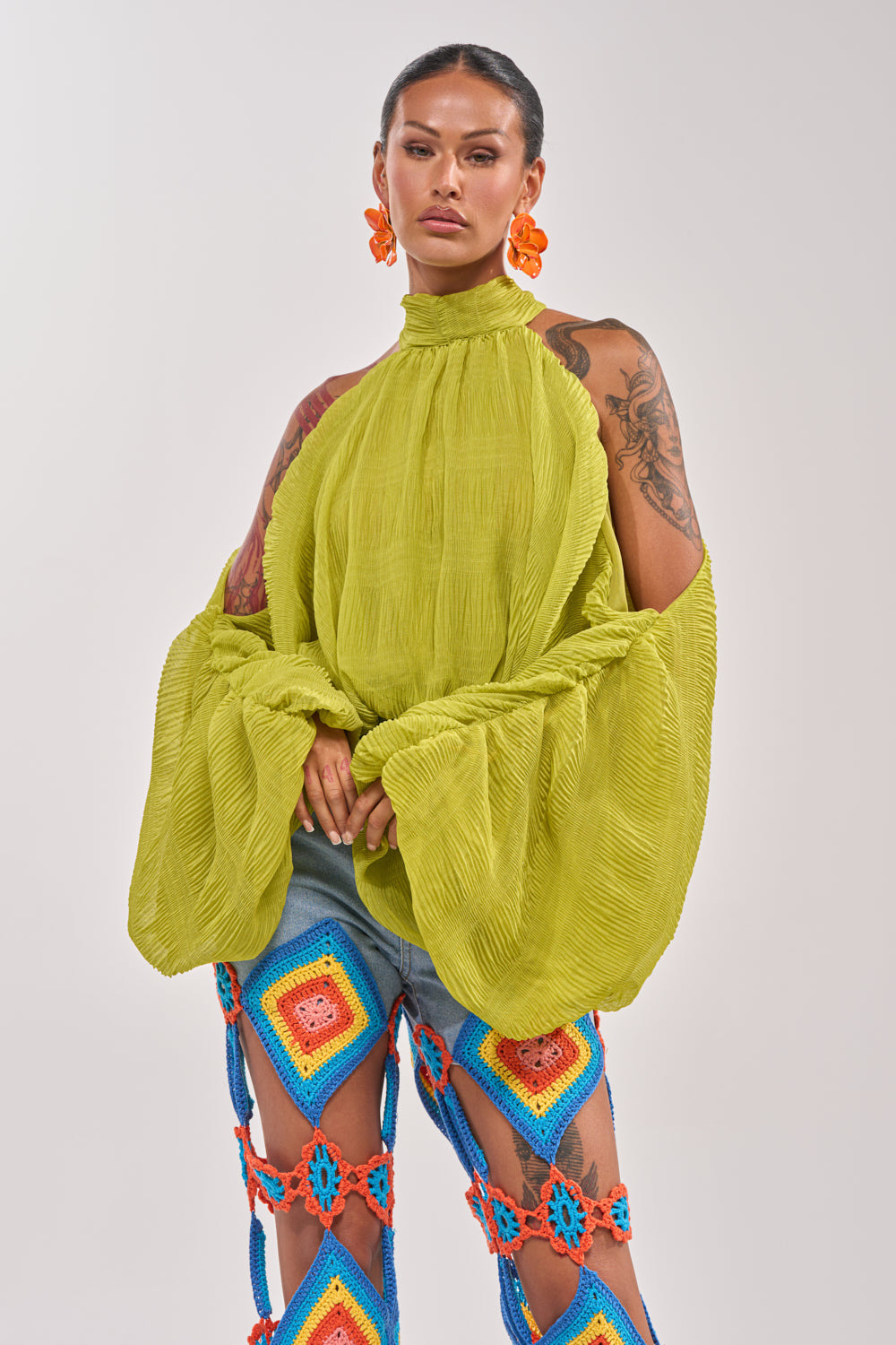 A person with long dark hair wears the NOT PLAYING BLOUSE in lime green with balloon sleeves and a high neck, paired with colorful open-patterned crochet pants and bold orange floral earrings, posing against a light background. 0