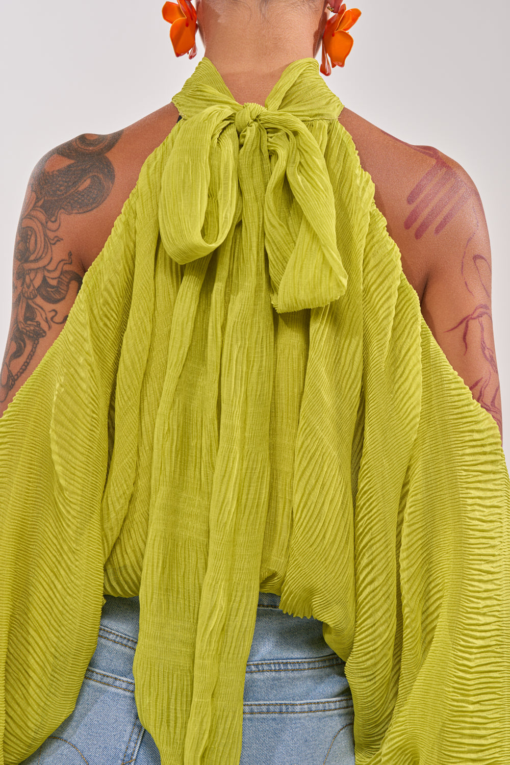 A person wears the NOT PLAYING BLOUSE, a bright green chiffon halter cold shoulder top with pleats and a large back bow, paired with blue jeans and orange flower earrings. Flowing fabric and exposed shoulders reveal visible tattoos. 9