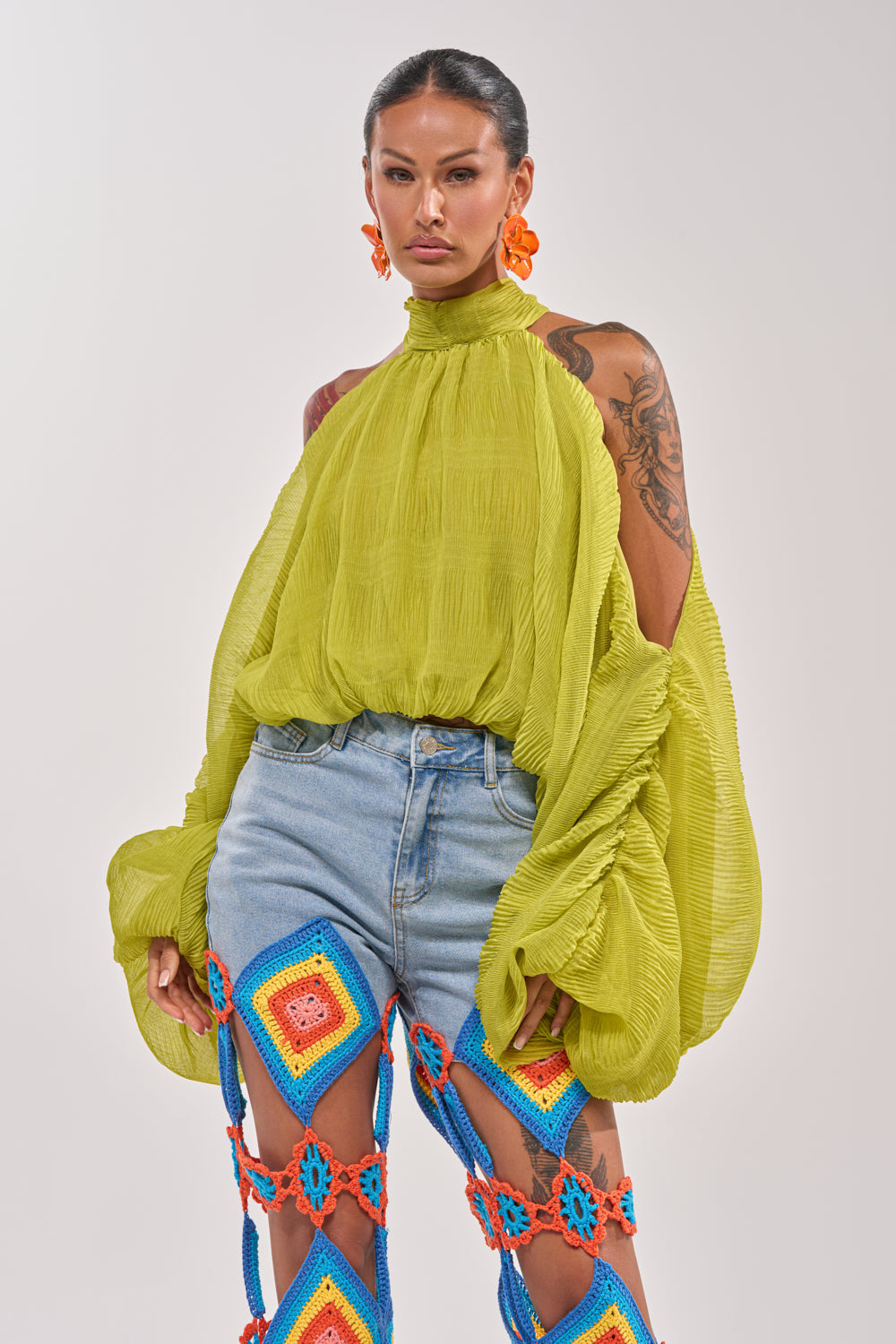 A woman wears the NOT PLAYING BLOUSE, a loose light green chiffon top with dramatic balloon sleeves, paired with light blue jeans featuring colorful crocheted cutouts and bright orange earrings against a plain background. 1