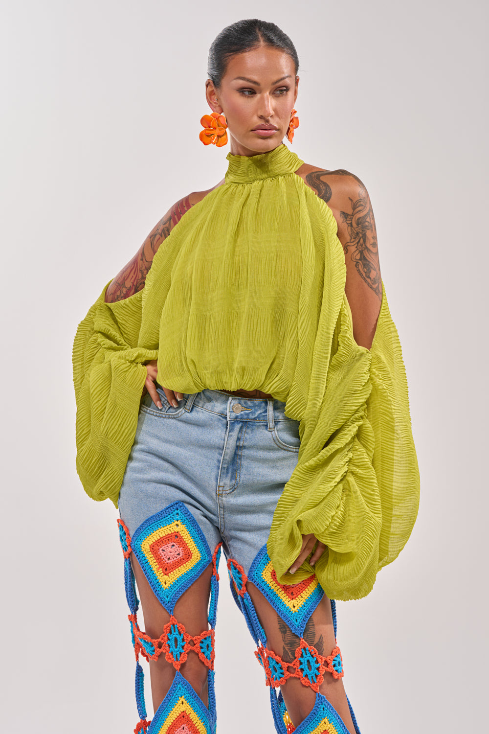 A woman poses confidently in the NOT PLAYING BLOUSE, a light green chiffon top with balloon sleeves, paired with colorful crochet pants featuring open sections. She accessorizes with bold orange flower earrings and displays arm tattoos. 2